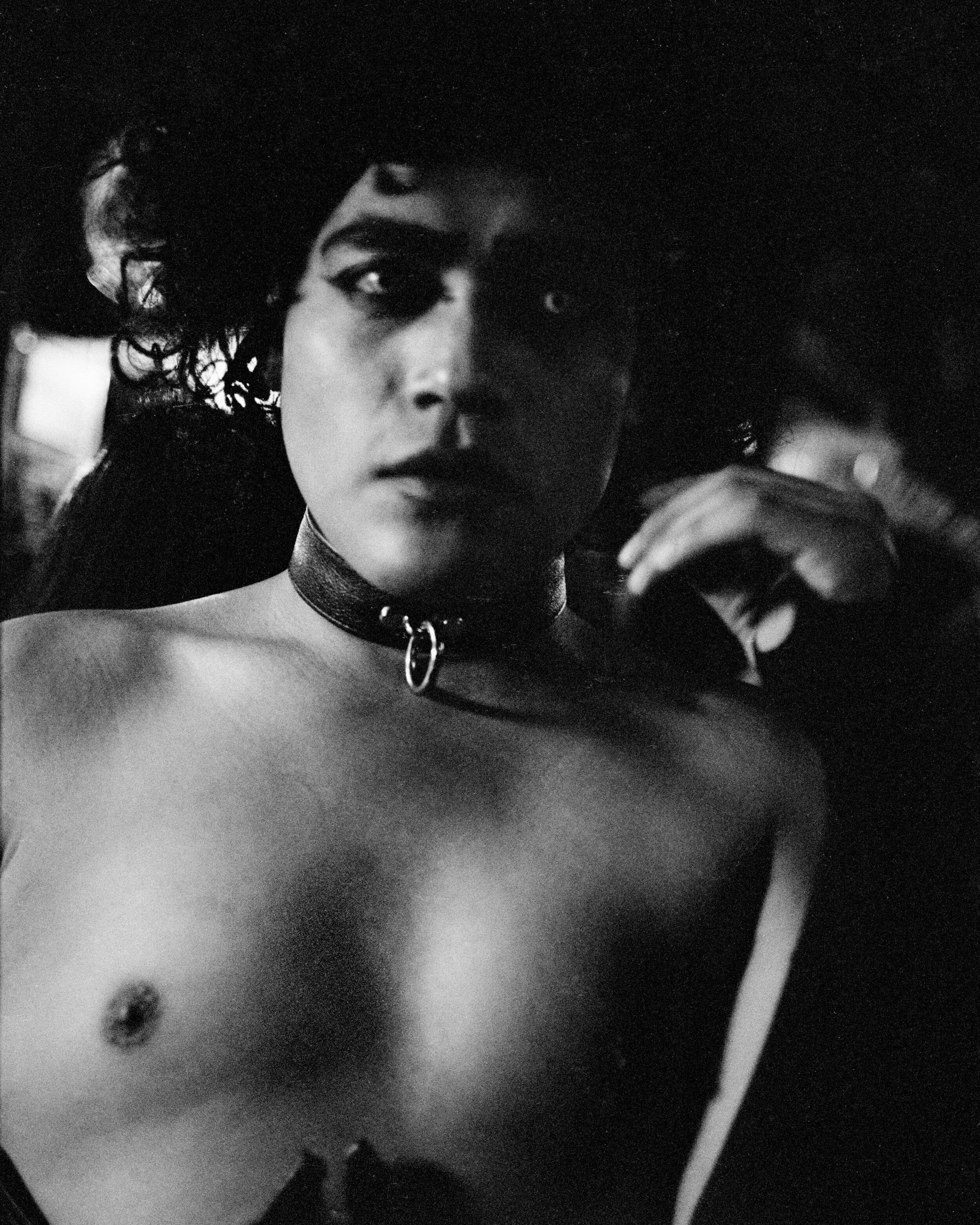 Black and white photo of a person with dark curly hair, wearing a choker with a ring, looking directly at the camera, with an exposed shoulder and hand touching their face.