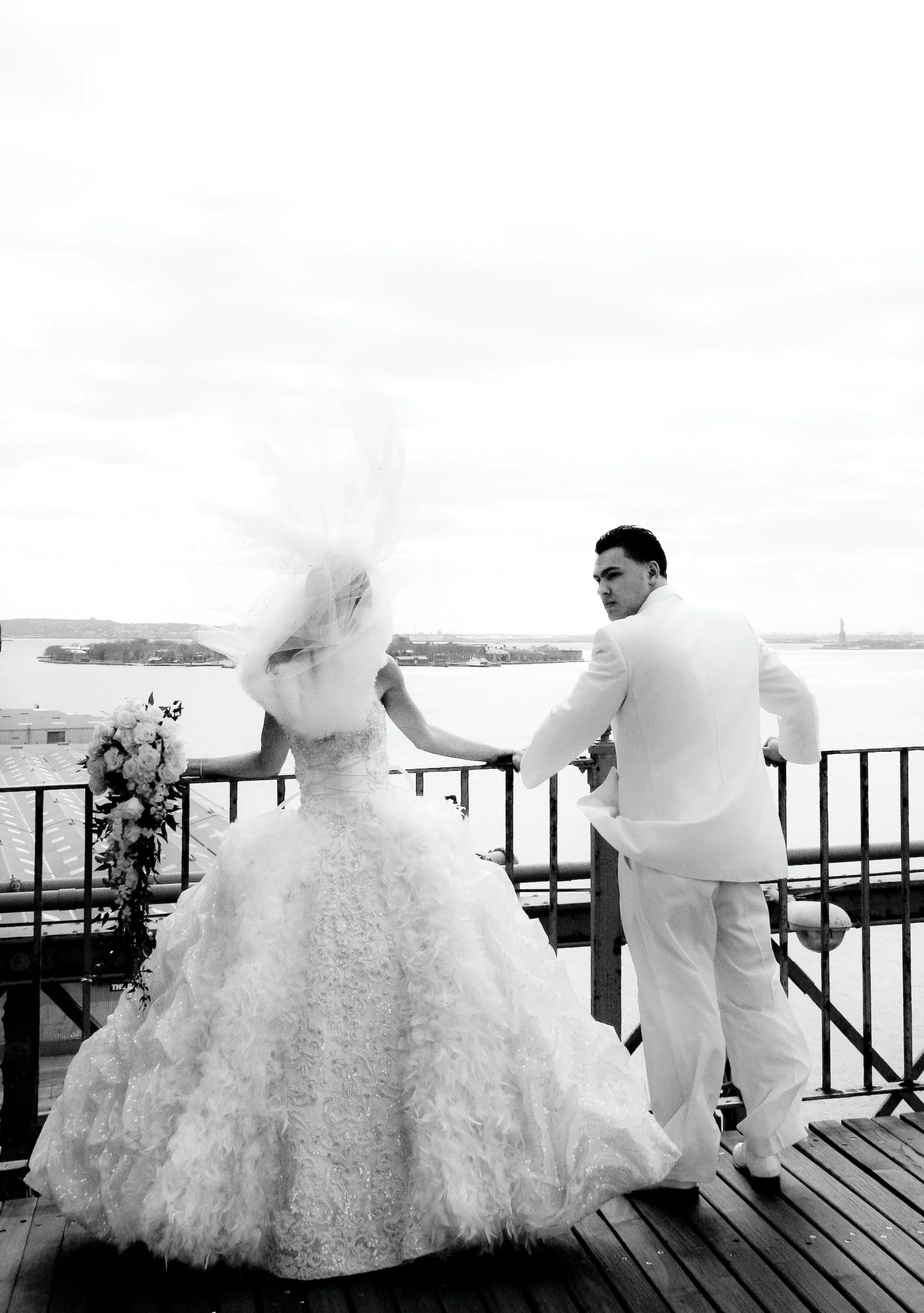 Black and white photo of a bride and groom standing on a balcony holding hands, overlooking a body of water with a city skyline in the distance.