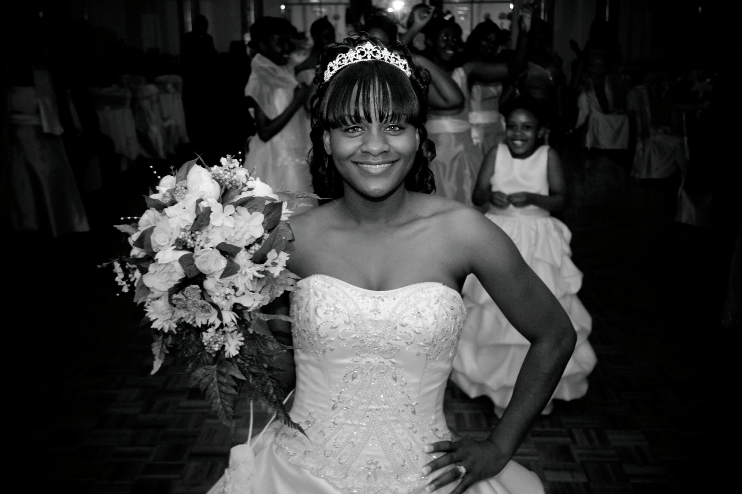 A bride in a strapless wedding gown with beadwork, wearing a tiara, smiling, holding a bouquet of flowers, and posing indoors at a wedding reception with children and guests in the background.