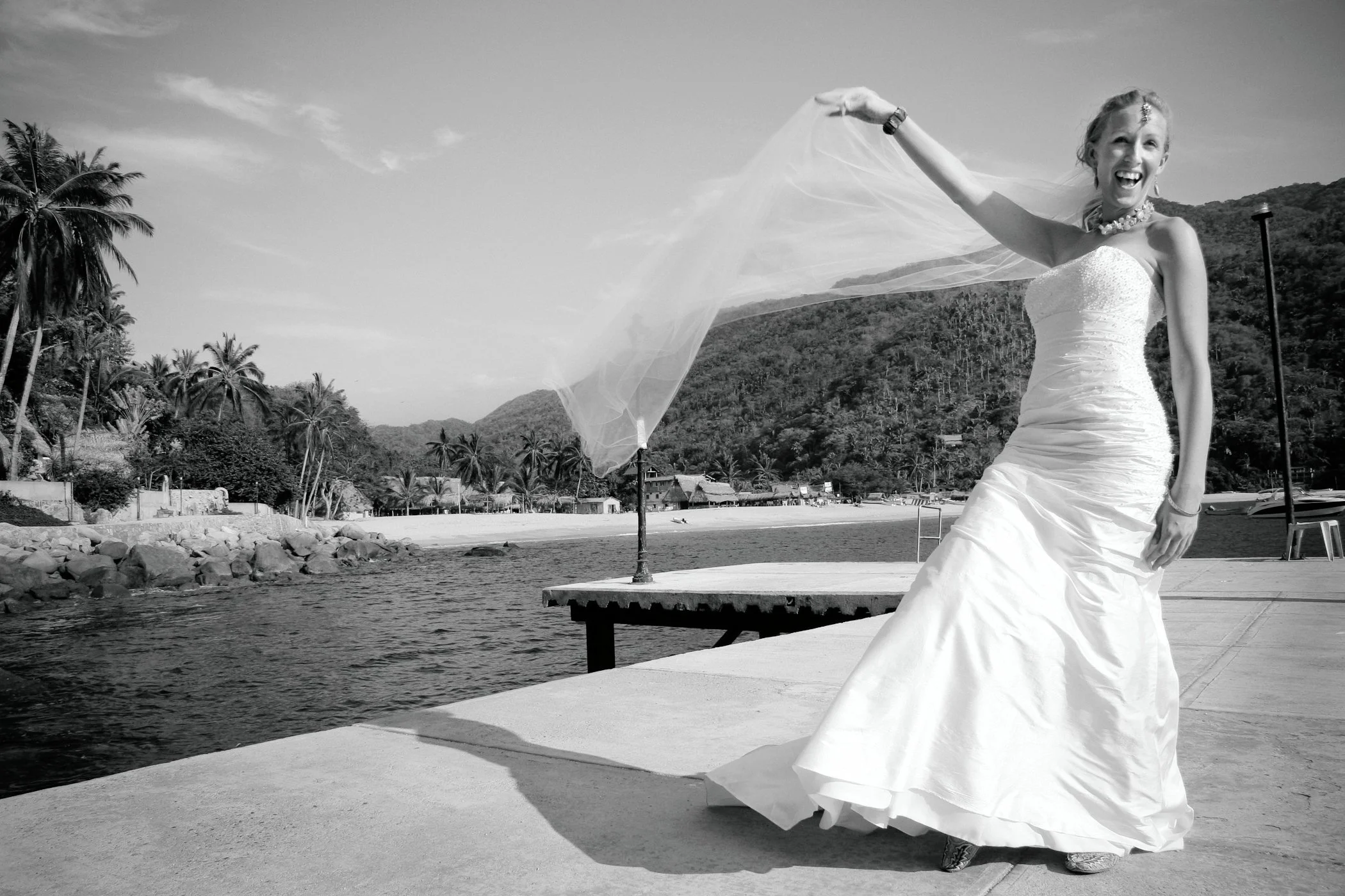 A woman in a wedding dress standing on a dock near water, smiling and holding her veil in the air with hills and palm trees in the background.