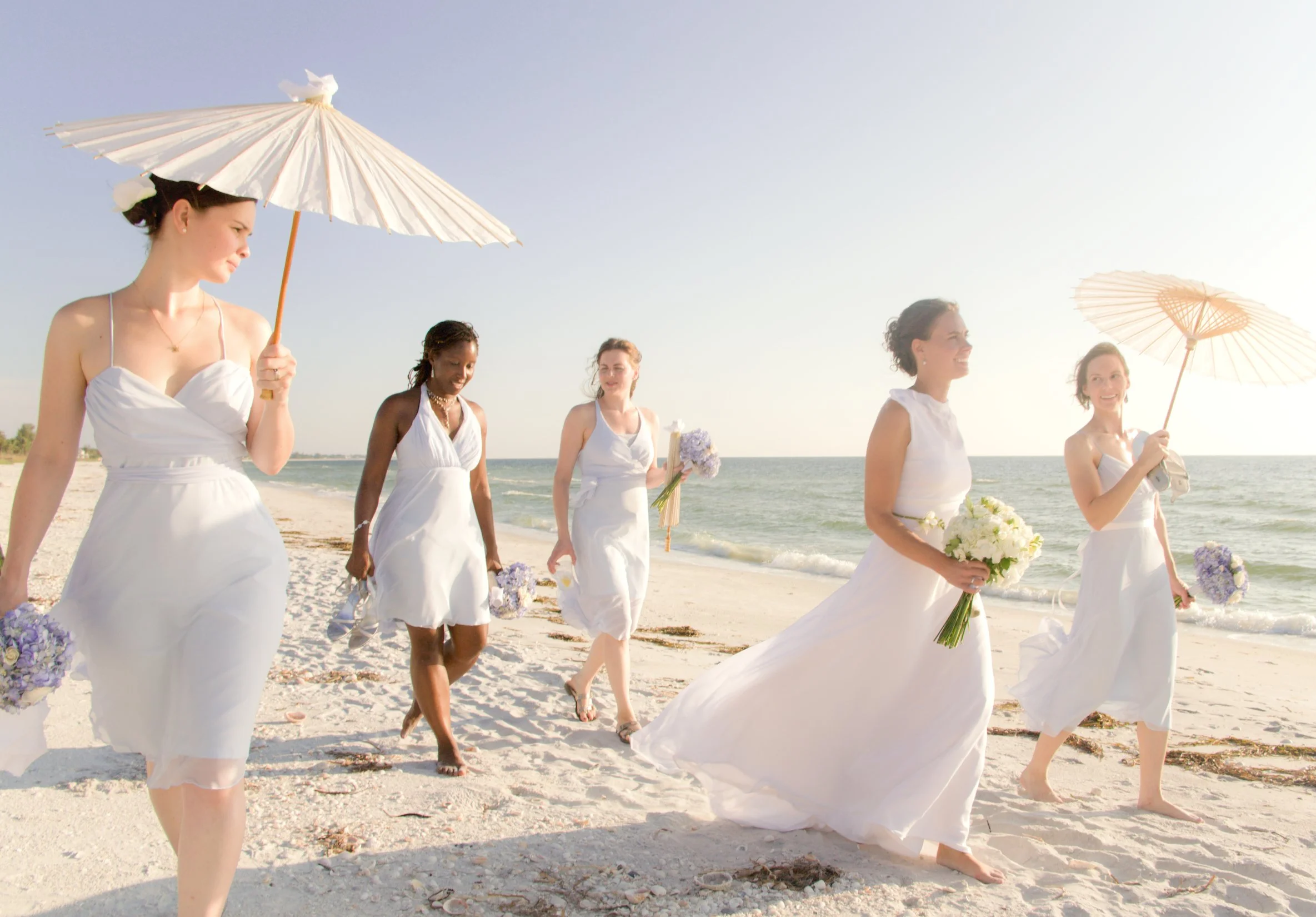 A group of women in white dresses walking on a beach, some carrying umbrellas and flowers, during a sunny day.
