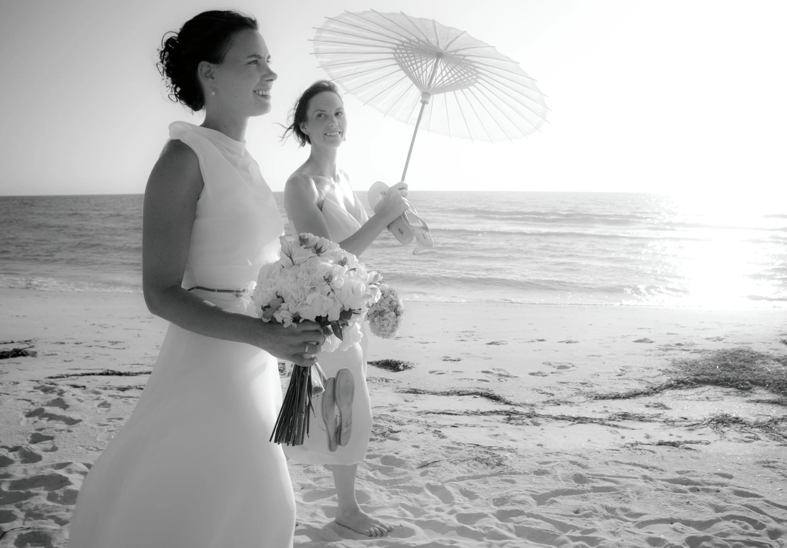 Two women in wedding dresses walking on the beach, one holding a bouquet and the other holding an umbrella, with the ocean and a bright sky in the background.