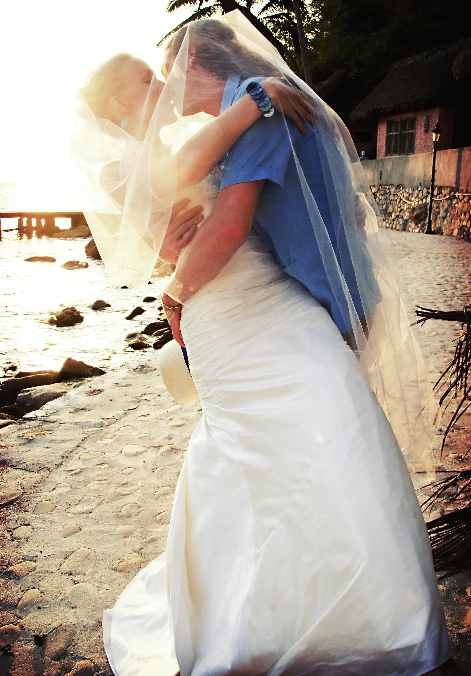 A woman in a wedding dress with a veil, hugging a person in a blue shirt on a beach during sunset.