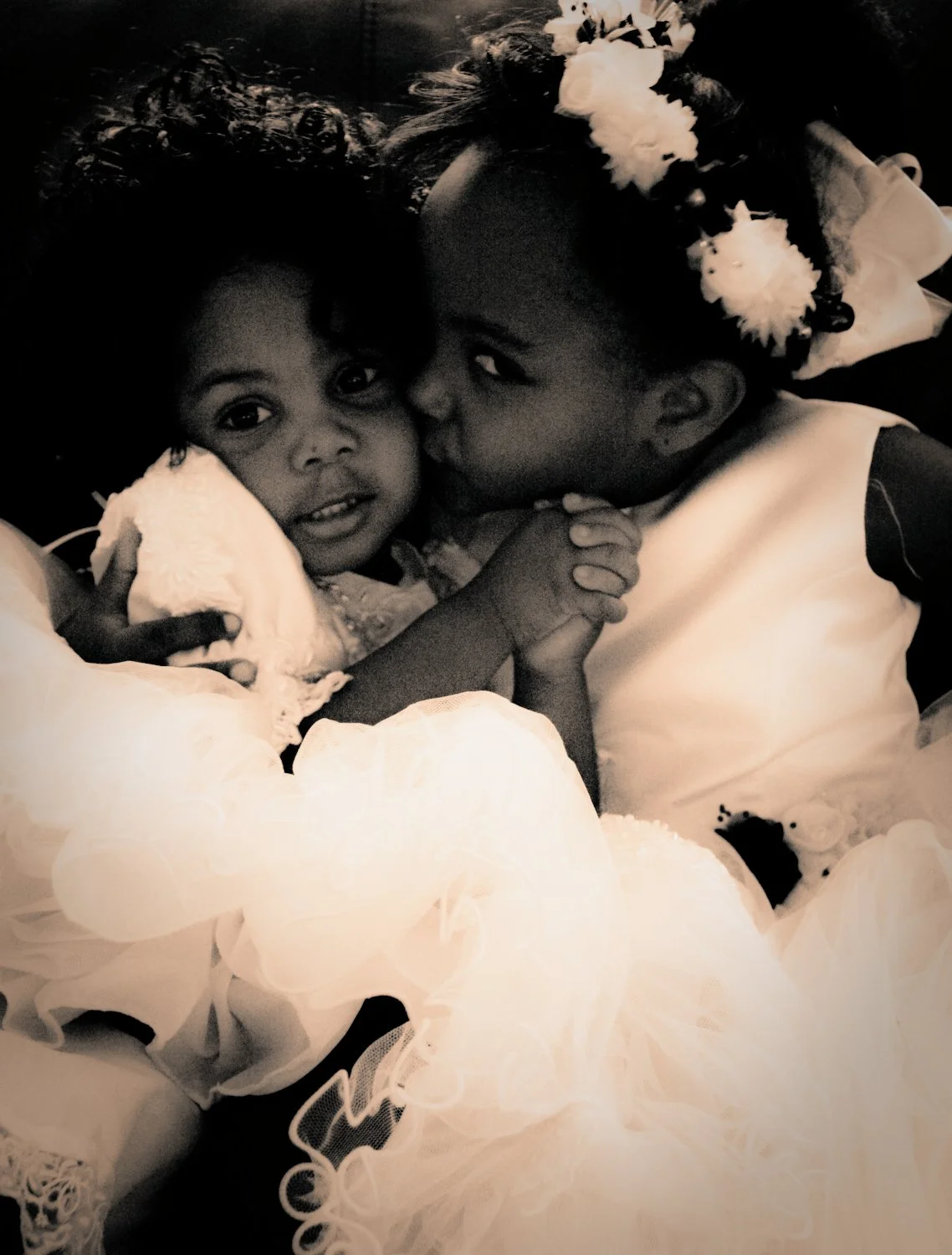 Two young girls with dark hair, one kissing the other on the cheek, wearing fancy dresses and floral accessories in their hair, lying close together, holding hands, on a dark background.