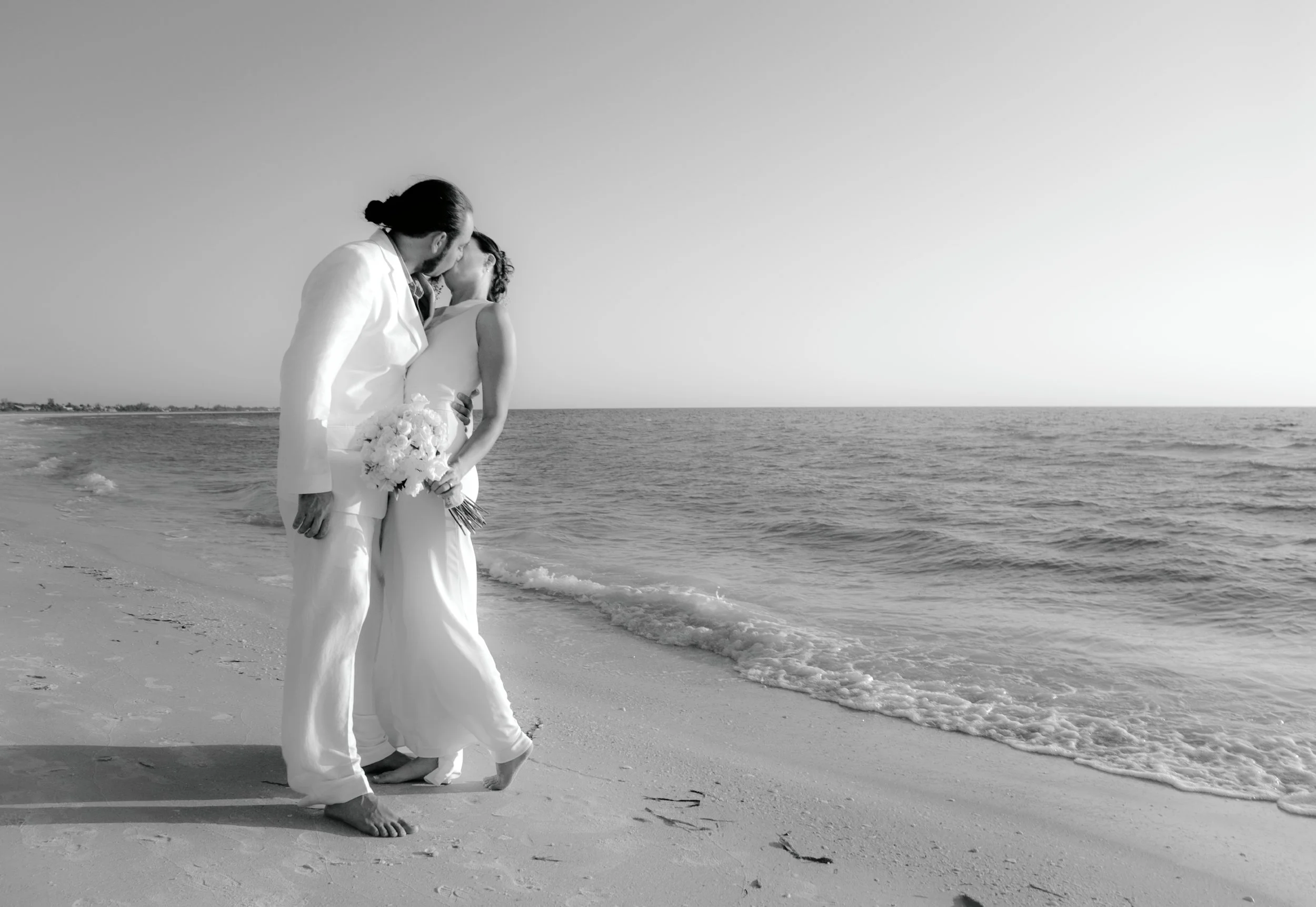 A black-and-white photo of a couple in wedding attire sharing a kiss on the beach, with the ocean and sky in the background.