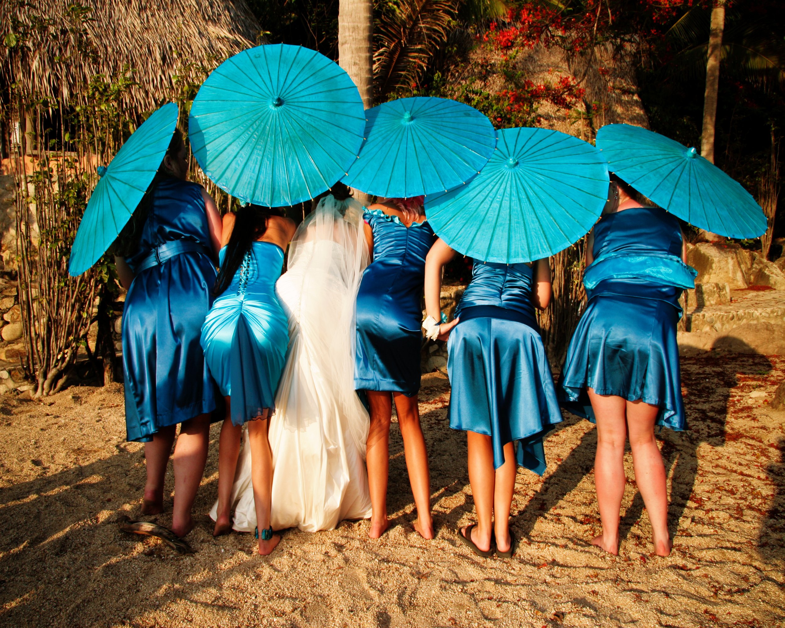 A group of women dressed in matching blue dresses and to the same height, holding blue umbrellas, standing on sandy ground with tropical plants in the background.