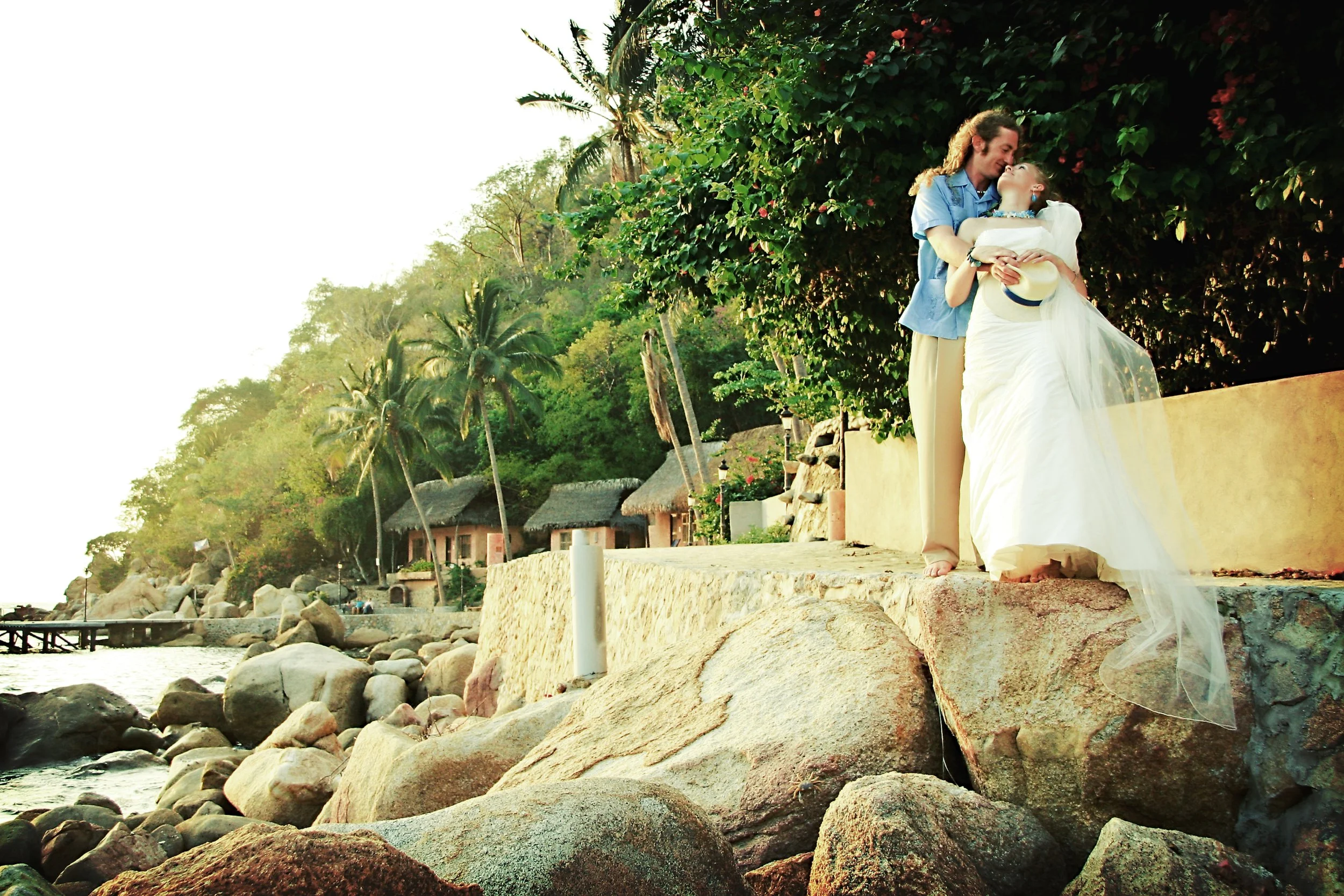 A man and a woman are standing on a rocky shoreline, embracing each other. The woman is dressed in a white wedding gown and holding a hat, while the man is wearing a blue shirt and khaki pants. Behind them, there are lush green trees, including palm 