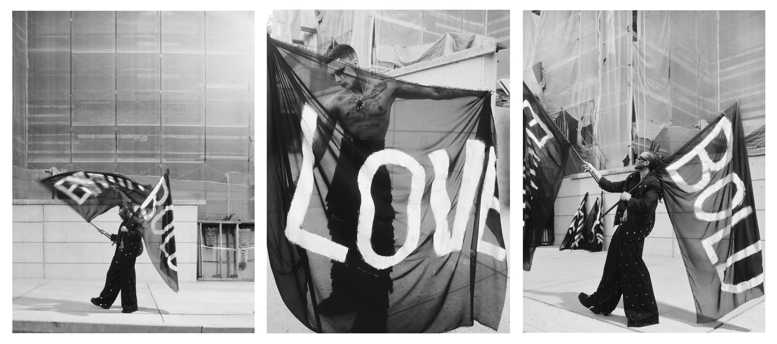 A man with face paint and dreadlocks, dressed in a dark outfit, holds a flag and performs during a street protest or demonstration. The scene is captured in black and white, with a background of a building and construction scaffolding.