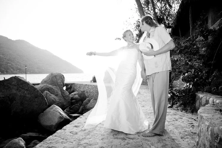A woman in a wedding dress and a man in casual clothes stand on a rocky beach, smiling and looking at each other, with a lake and mountains in the background.
