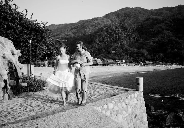 A woman in a dress and a man walking along a beachside promenade with mountains in the background, the woman holding her dress as they walk.