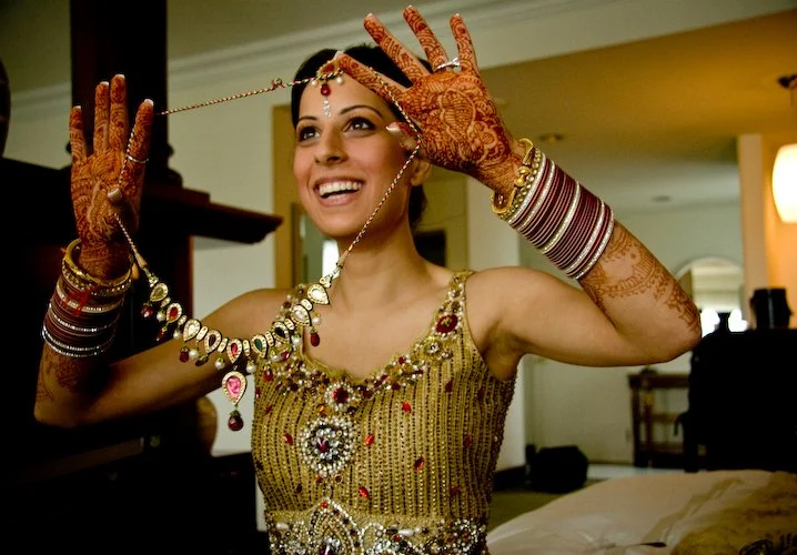 A smiling woman wearing traditional Indian jewelry and clothing, holding a necklace with both hands. She has henna on her hands and arms, and the background appears to be a cozy indoor setting.