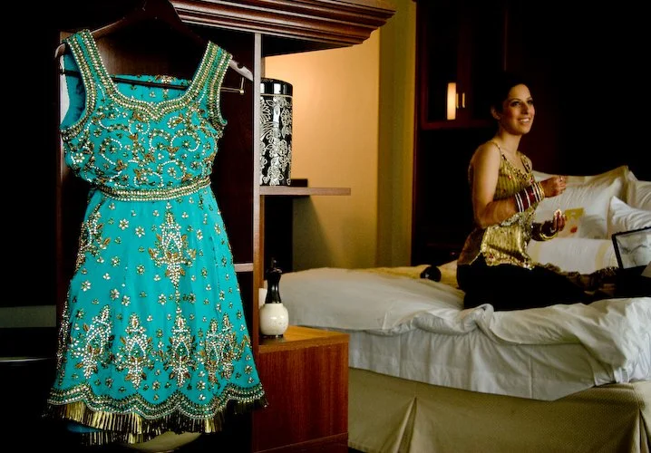 A turquoise traditional dress with intricate gold and silver embroidery hanging on a closet door in a hotel room. A woman sitting on the bed in a matching outfit, smiling and looking towards the camera.