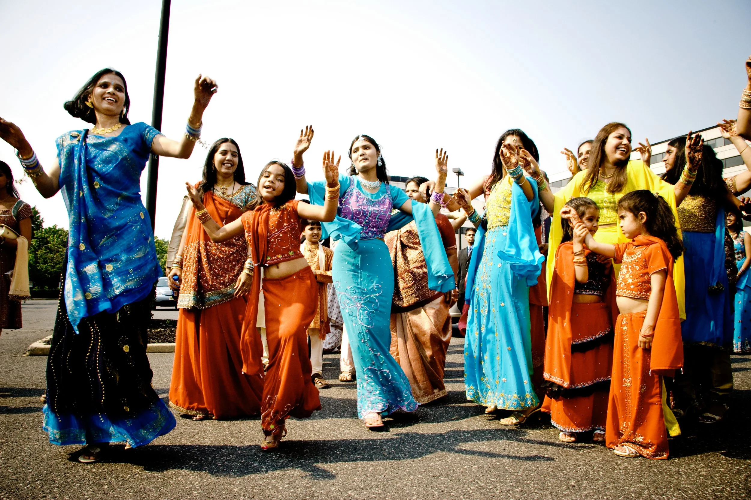 Group of women and children of Indian ethnicity dancing outdoors in colorful traditional clothing.