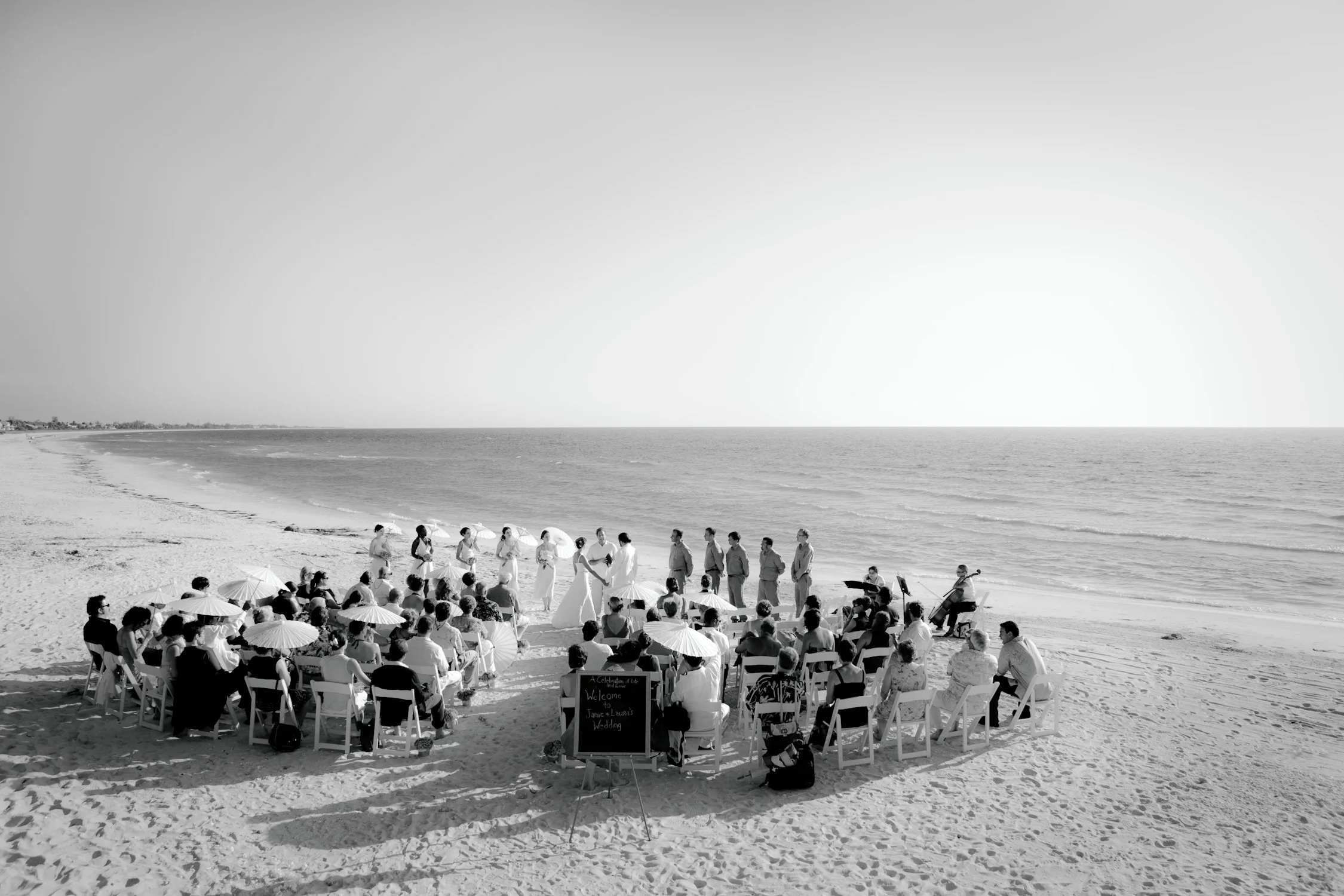 Black and white photo of an outdoor wedding ceremony on a beach, with guests seated in chairs holding umbrellas, facing the couple and officiant in front of the ocean.