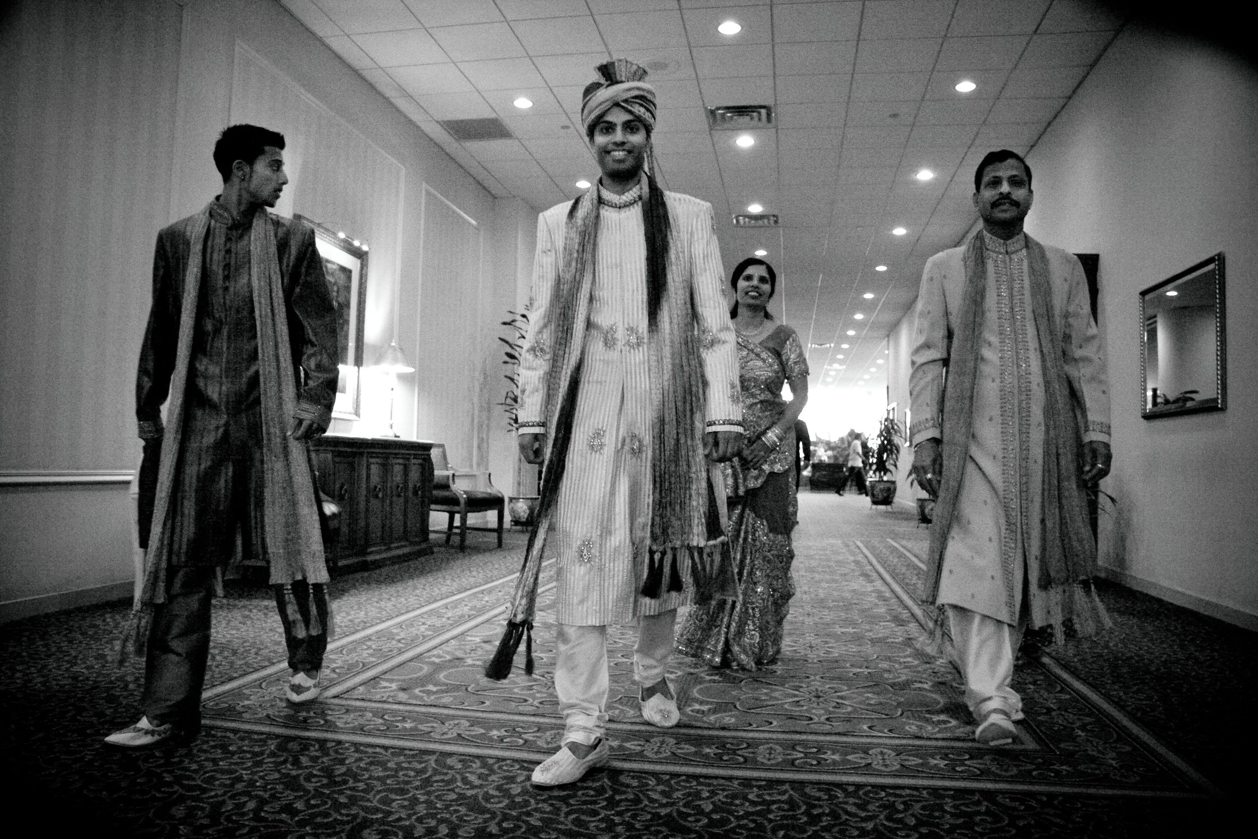 A group of five people walking down a hotel corridor, dressed in traditional Indian wedding attire. The central figure is a groom wearing a turban and sherwani, smiling at the camera. Behind him is a woman in a saree and three men in kurta and sherwa
