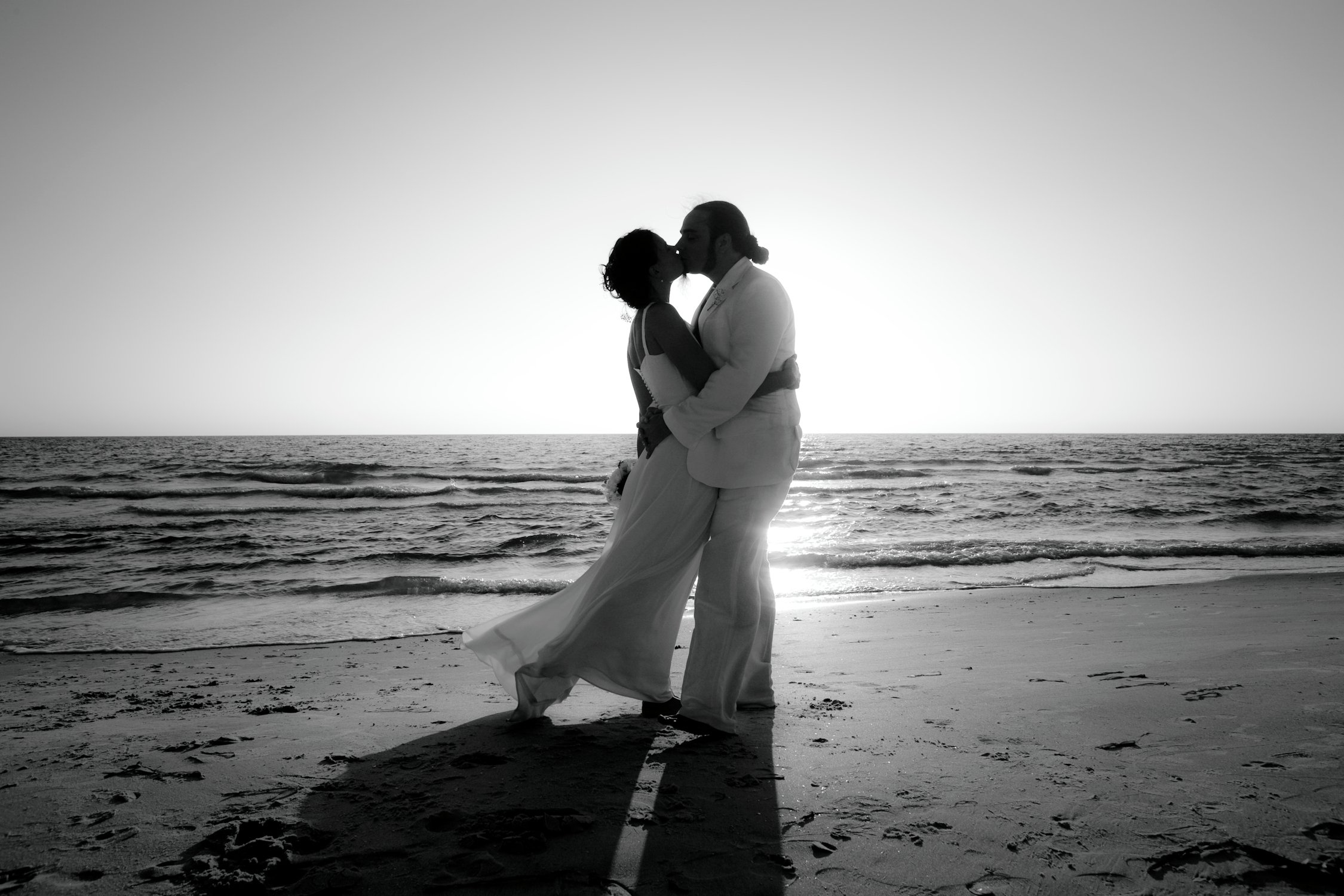 A couple dressed in wedding attire sharing a kiss on the beach at sunset, with the ocean and sky in the background.