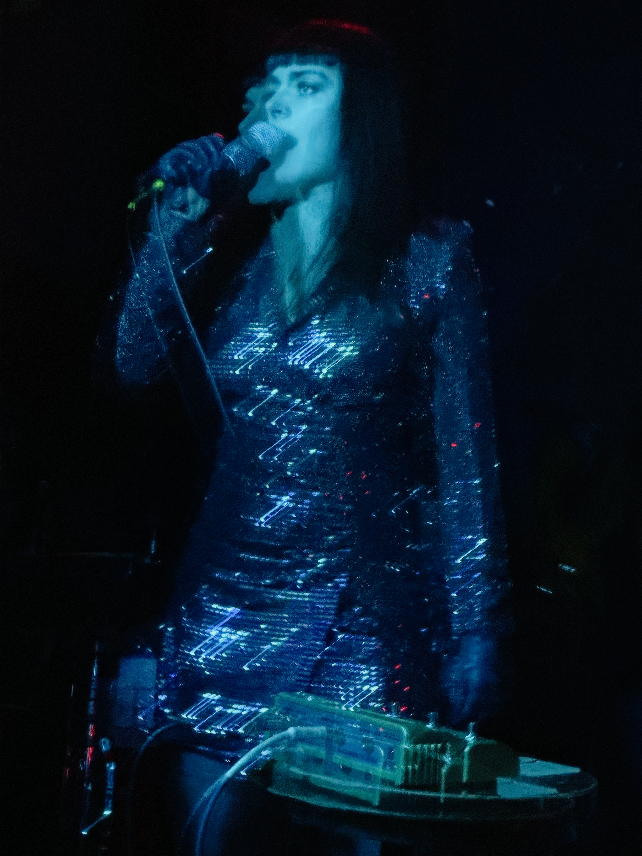 A woman with dark hair and bangs singing into a microphone on stage, illuminated by blue lighting, wearing a sparkly dress, with electronic music equipment in front of her.