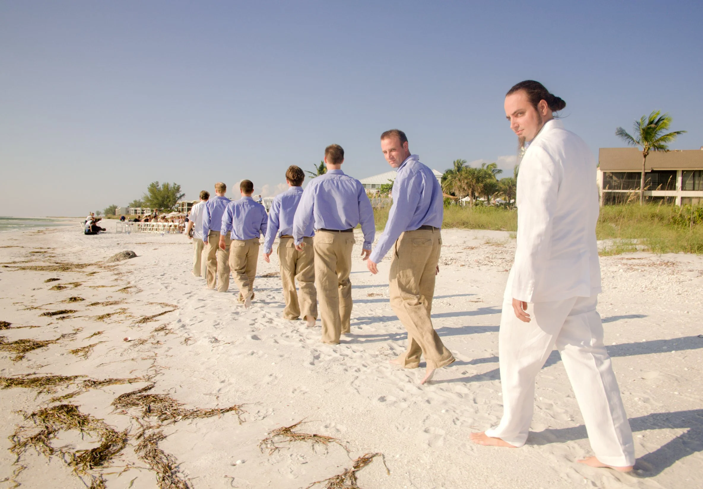 A group of men in dress shirts and beige pants walking along a sandy beach, with one man in white attire at the front, looking at the camera.