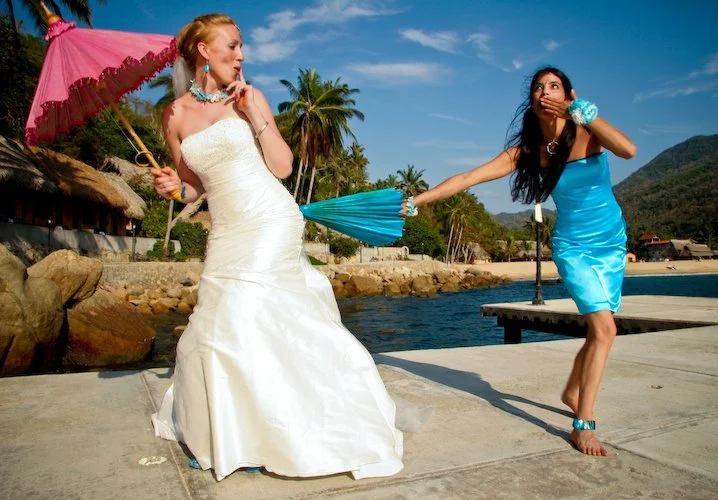 Two women in colorful dresses by a body of water, one holding a pink parasol and the other holding a blue parasol, in a tropical setting with palm trees and mountains in the background.