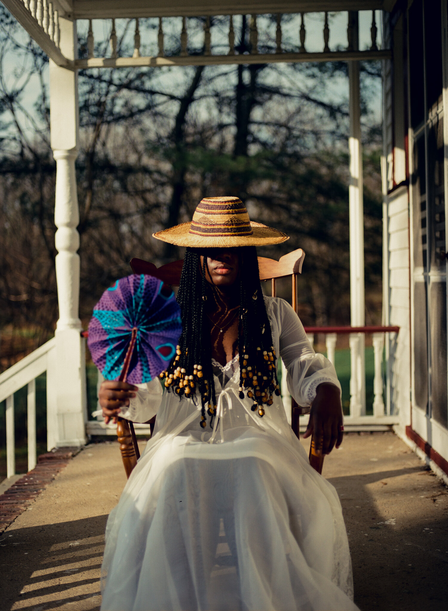 women with obscured face sits on porch holding purple fan in left hand