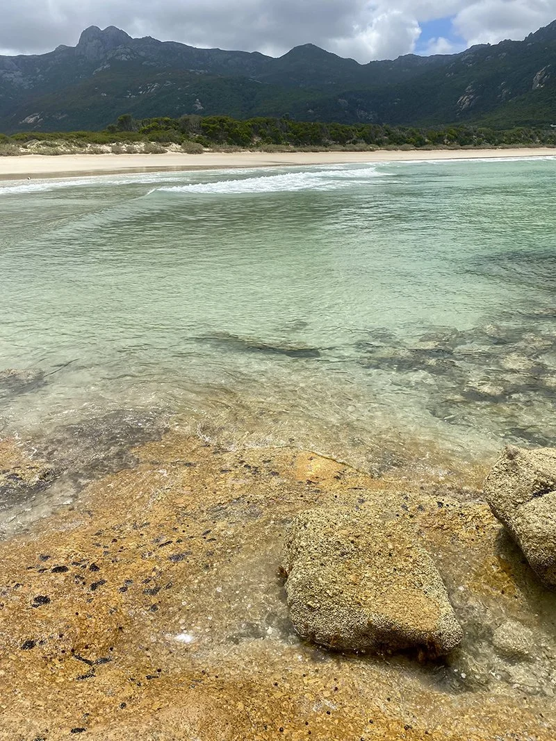 A beach scene with clear, shallow water over rocks in the foreground, a sandy shoreline in the middle, and green mountains with cloudy sky in the background.