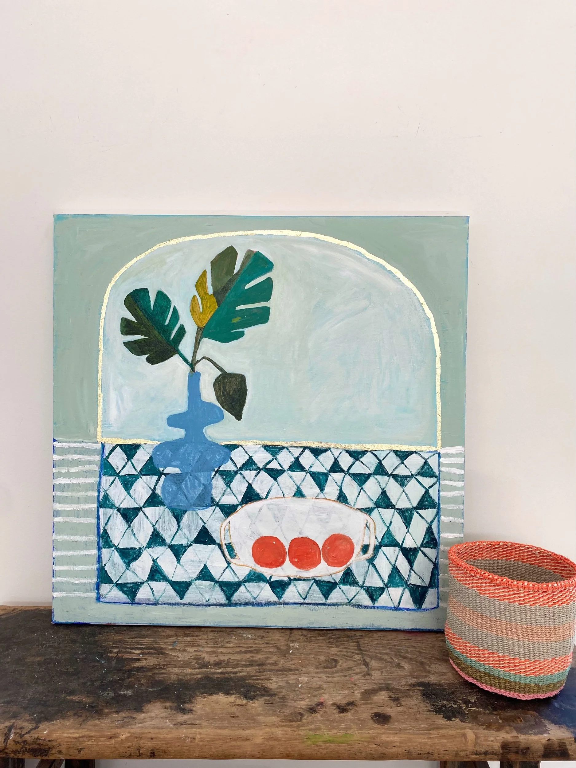 Painting of still life on table with a vase holding a plant and a bowl with three red balls. Geometric patterned background. Basket next to the painting.