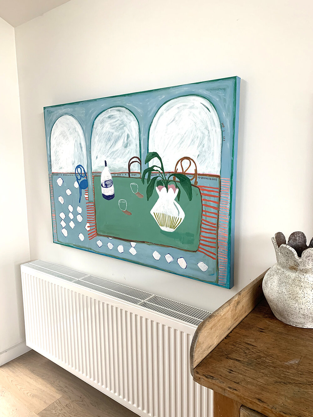 Painting on the wall depicting a table with assorted items and a plant in a vase, positioned above a white radiator next to a wooden surface and a ceramic vase.