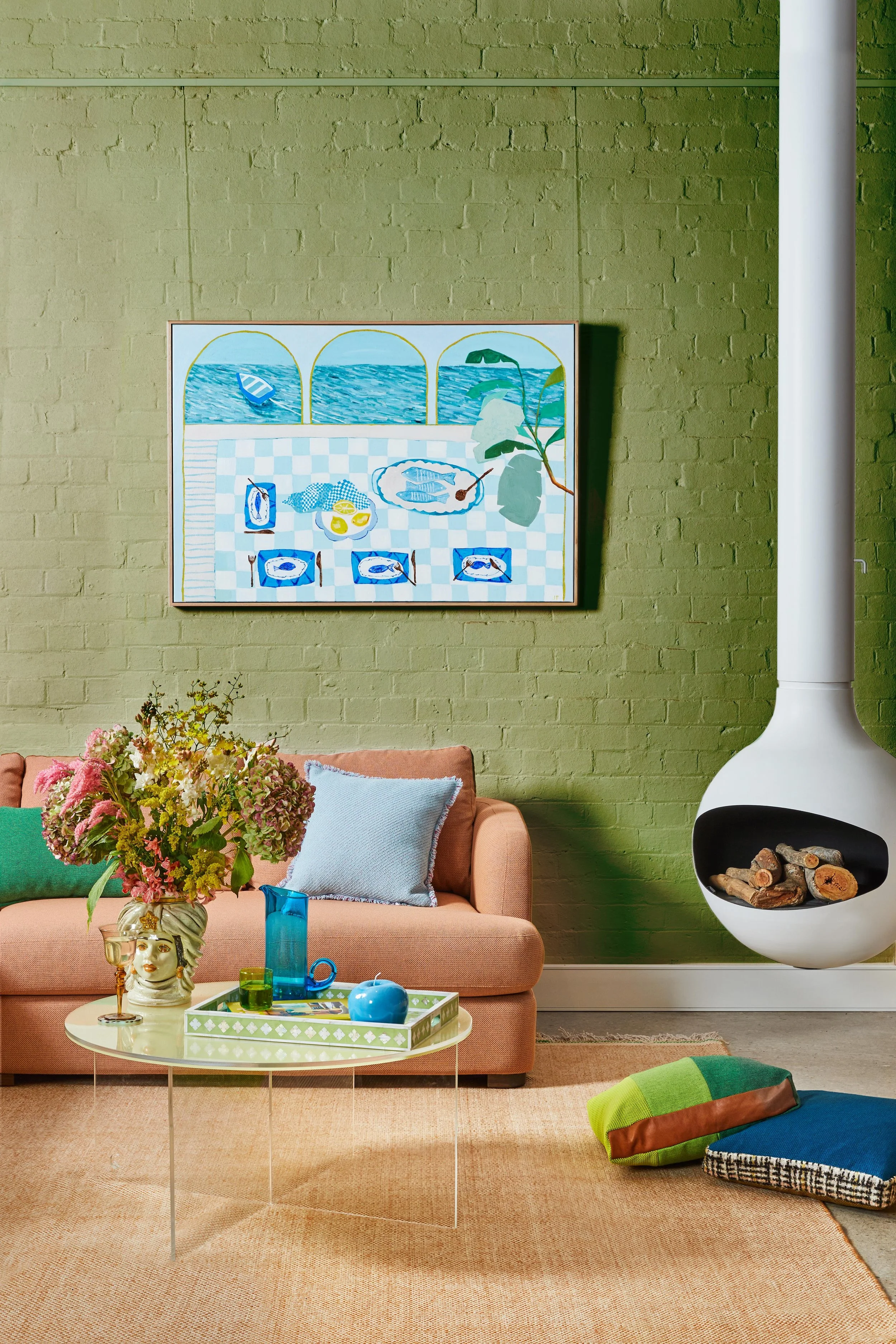 Interior of a cozy living room with a pastel peach sofa, colorful cushions, a modern round table with decorative items, and a green brick wall featuring a painting of a coastal scene. A white round fireplace is visible, and a rug lies on the floor.