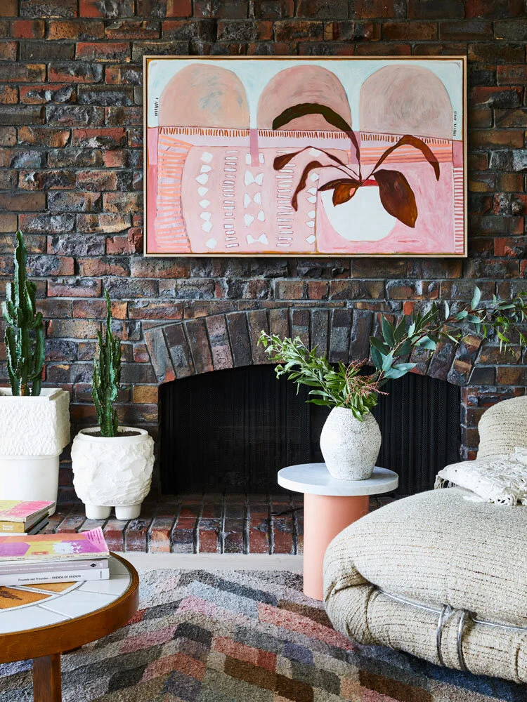 Interior with brick fireplace, abstract painting, potted plants, round marble side table, large cushioned chair, and colorful rug.