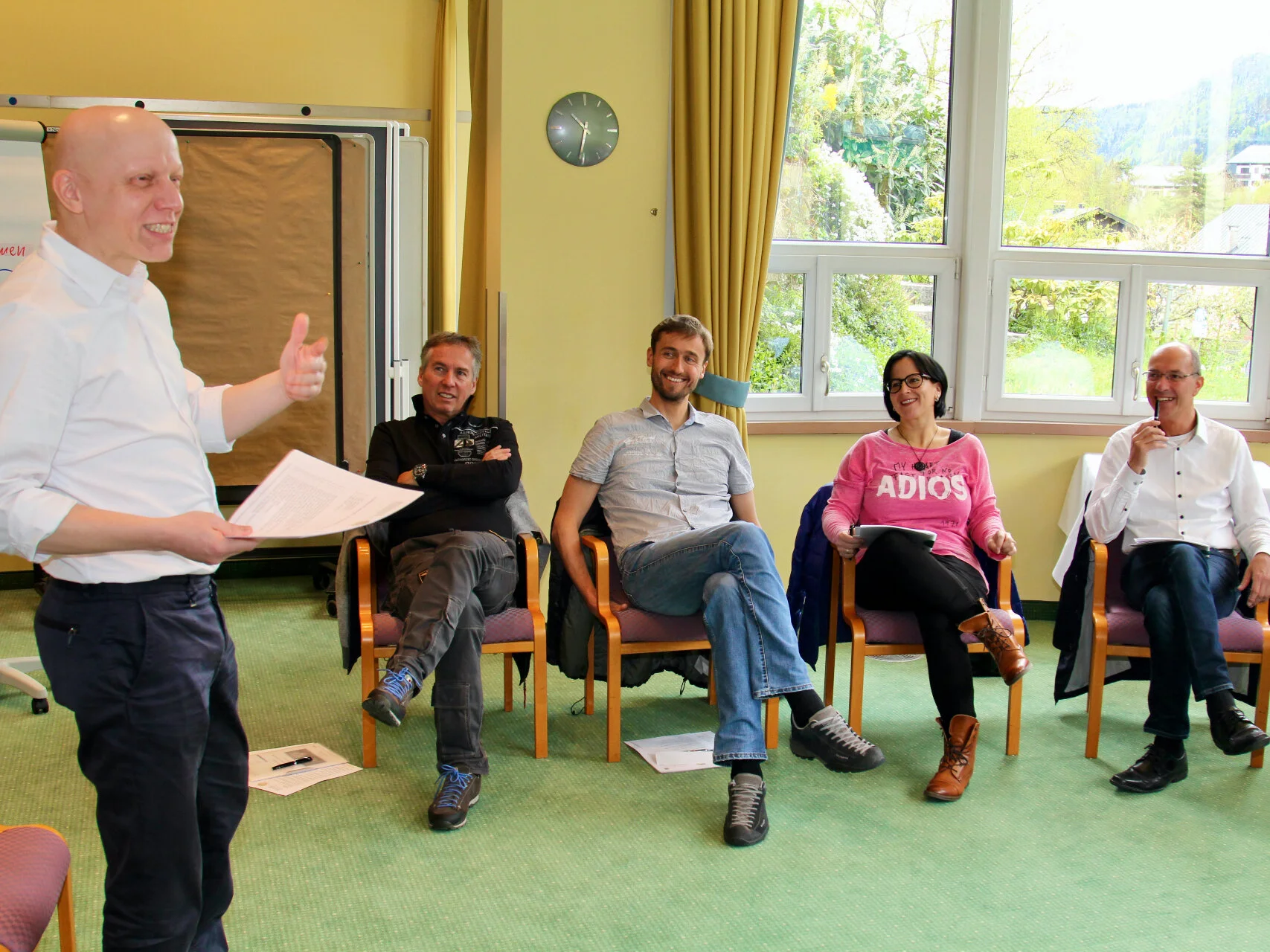 Lachender Mann in weissem Hemd steht vor mehreren sitzenden Menschen und spricht zu ihnen
