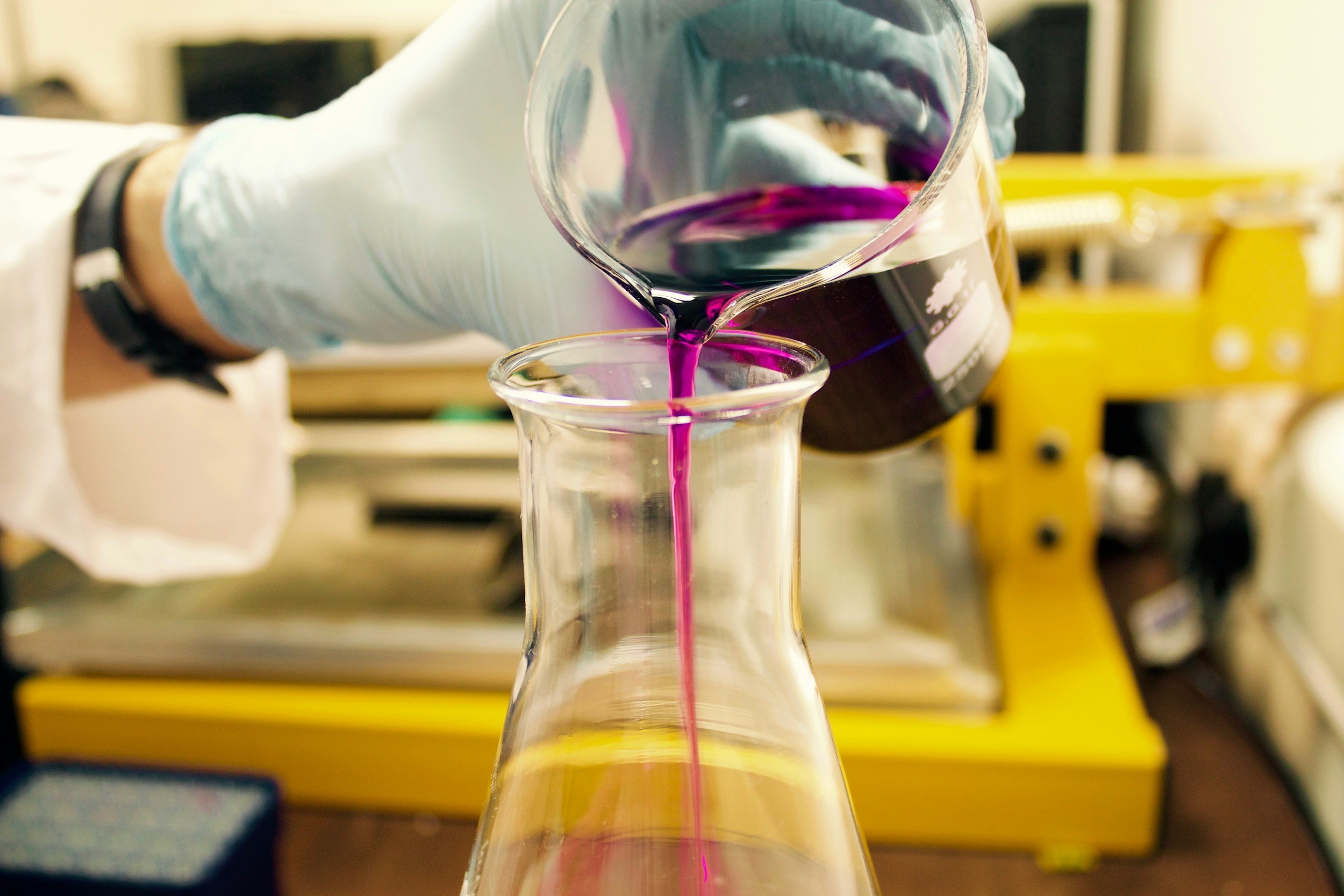 A person wearing a lab coat and gloves is pouring purple liquid into an Erlenmeyer flask in a laboratory setting.