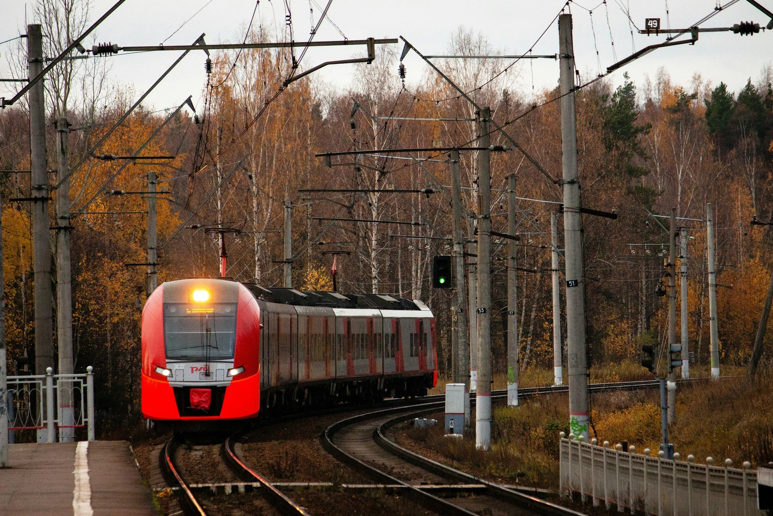 A modern red and gray train traveling on a curved railway track with open landscape and trees with fall foliage in the background. The train has a bright headlight and a green signal light is visible above the tracks.