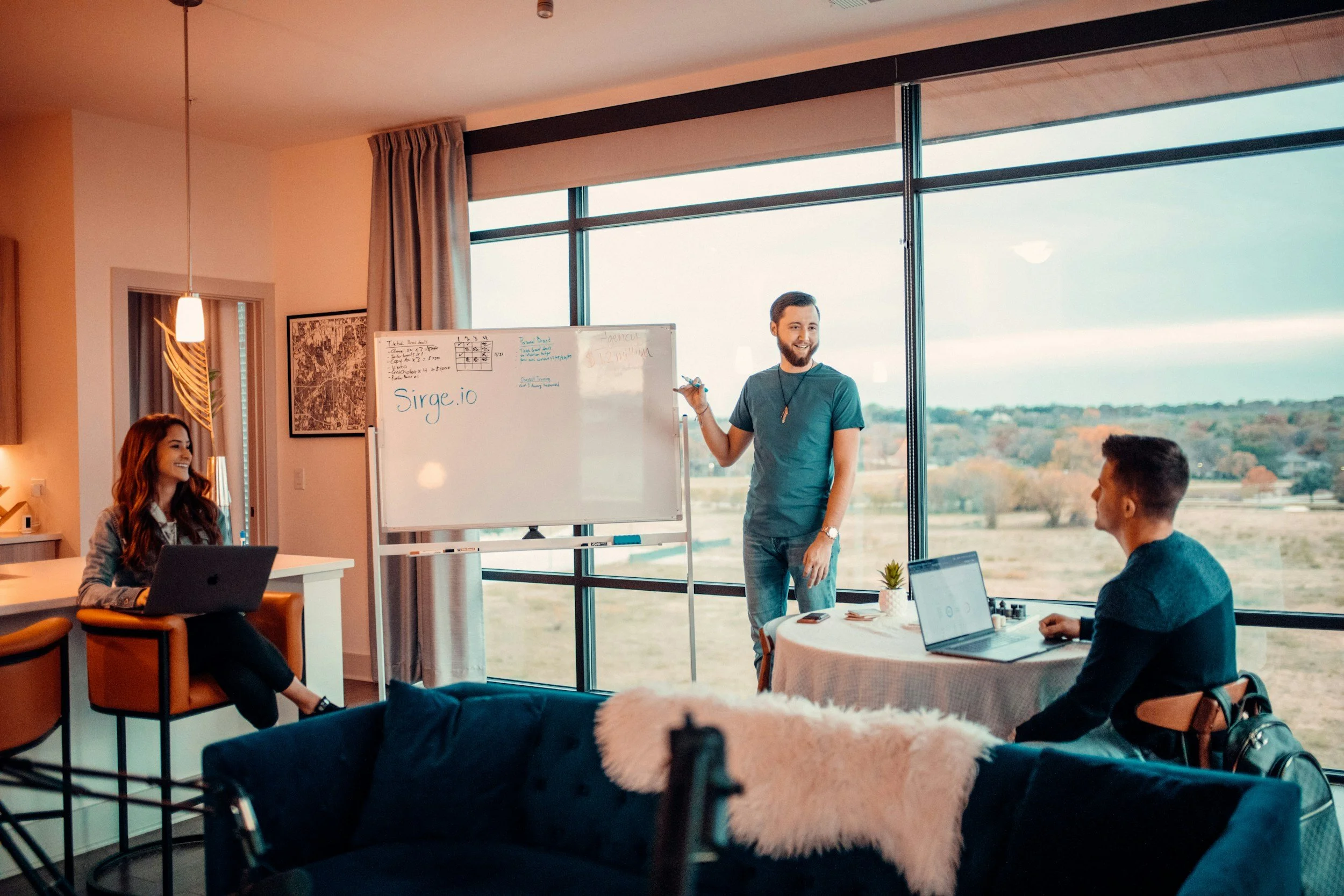 Three people in a meeting room with large windows overlooking a scenic landscape. One person is standing next to a whiteboard, speaking and holding a marker, while two others are seated, listening and working on laptops.