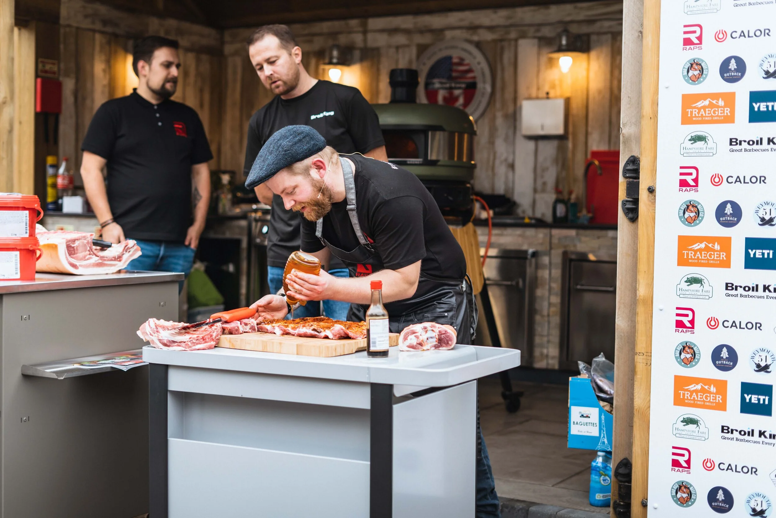 Three men in a rustic kitchen setting with one man applying sauce to meat on a cutting board, surrounded by raw cuts of meat and food preparation items, with brand logos displayed on the side.