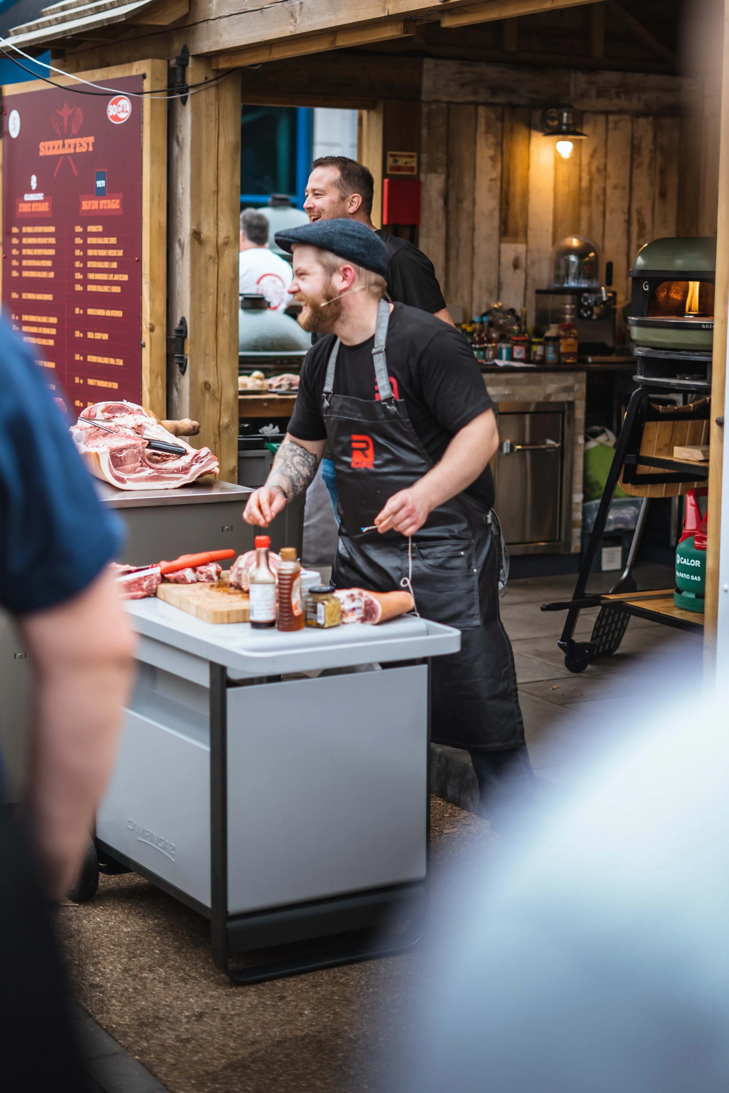 Man in apron at outdoor meat stall, preparing cuts of meat with various sauces and condiments on the table.