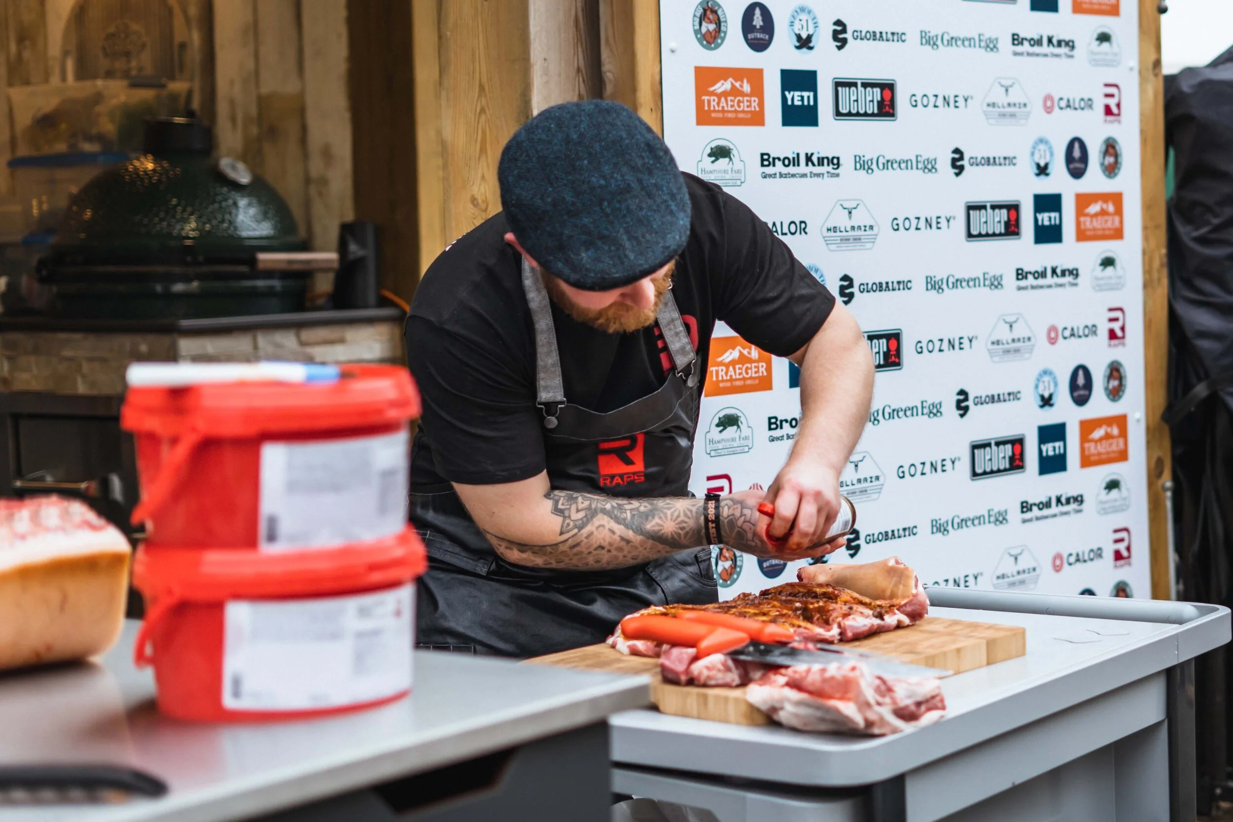 A person seasoning meat at a grilling event with brand logos in the background.