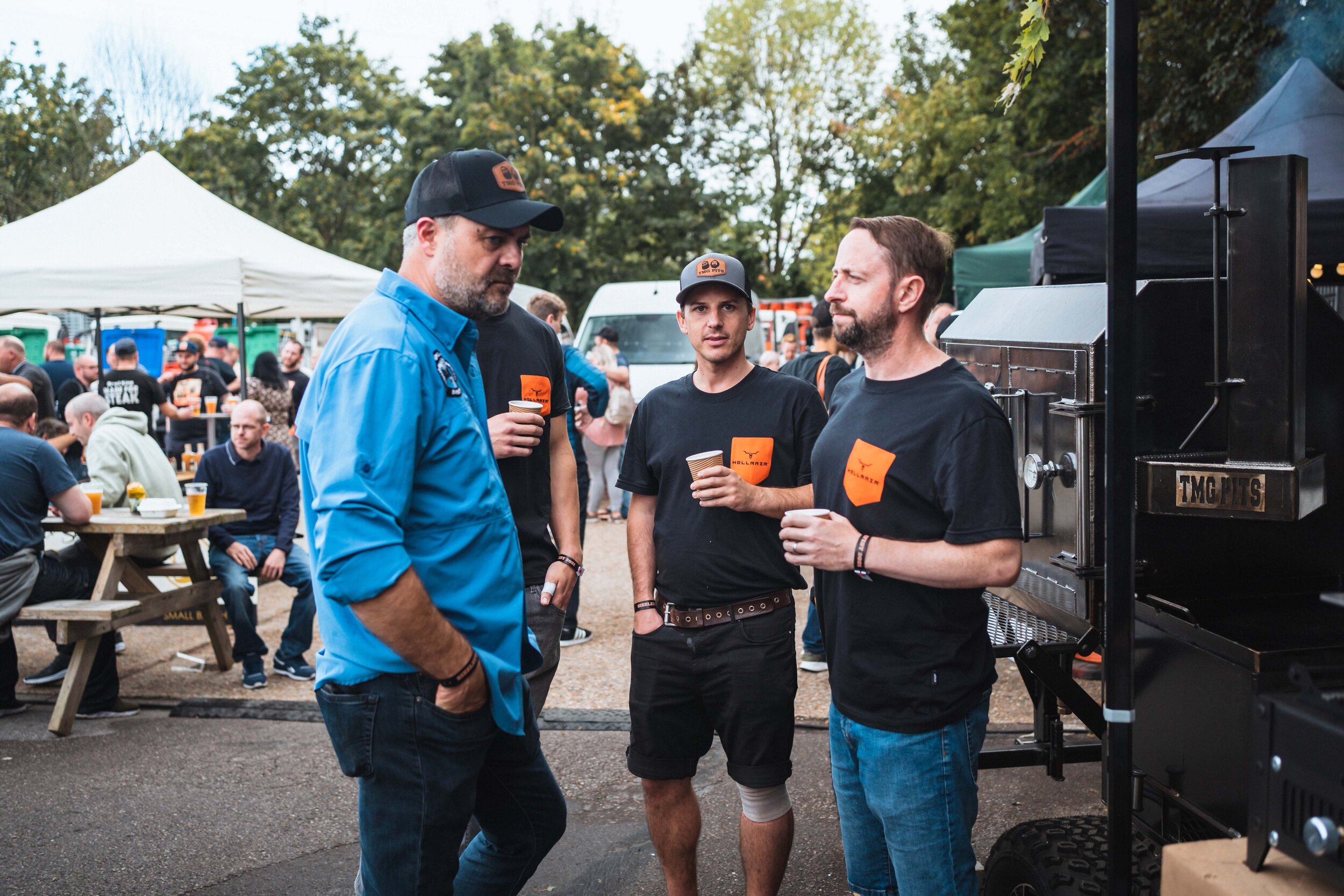 Outdoor barbecue event with people socializing and holding drinks, a smoker grill labeled "TMC Pits" in the foreground.