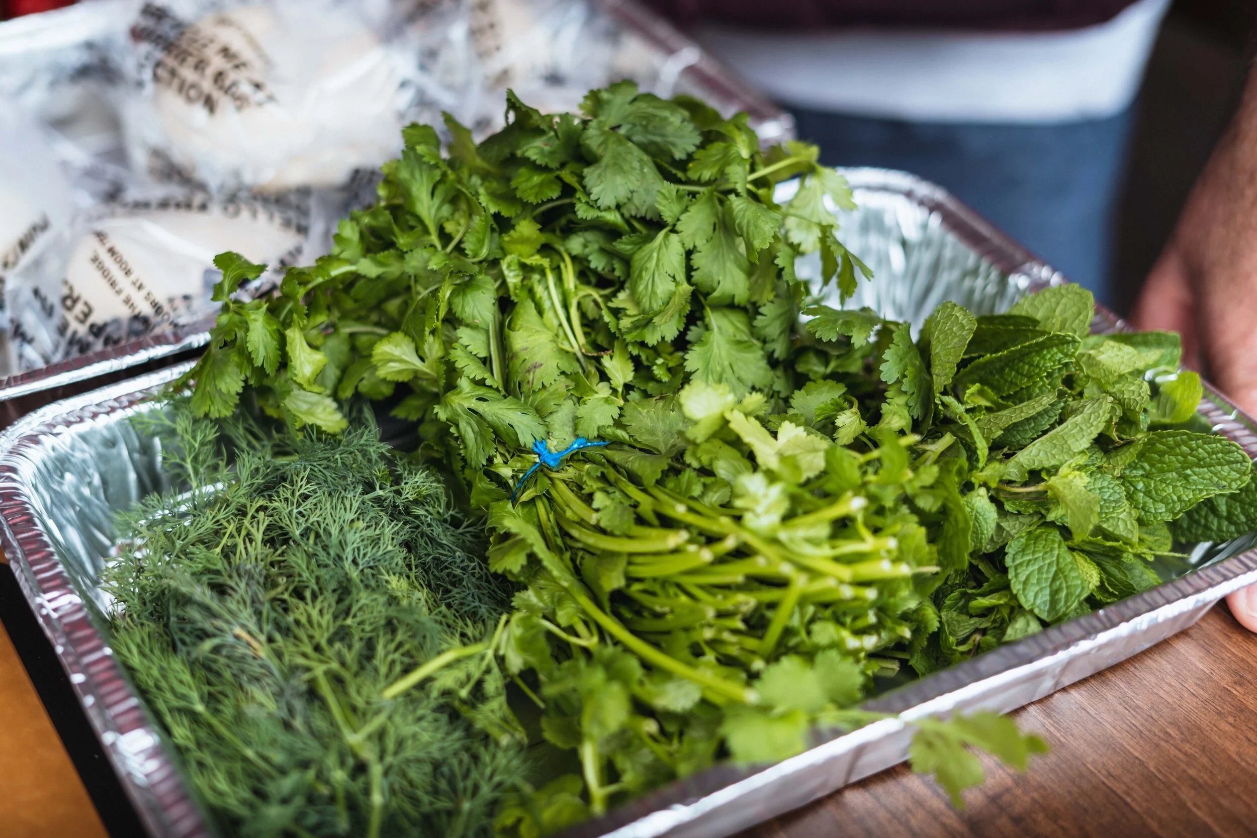 Fresh herbs including cilantro, dill, and mint in a metal tray on a wooden surface.
