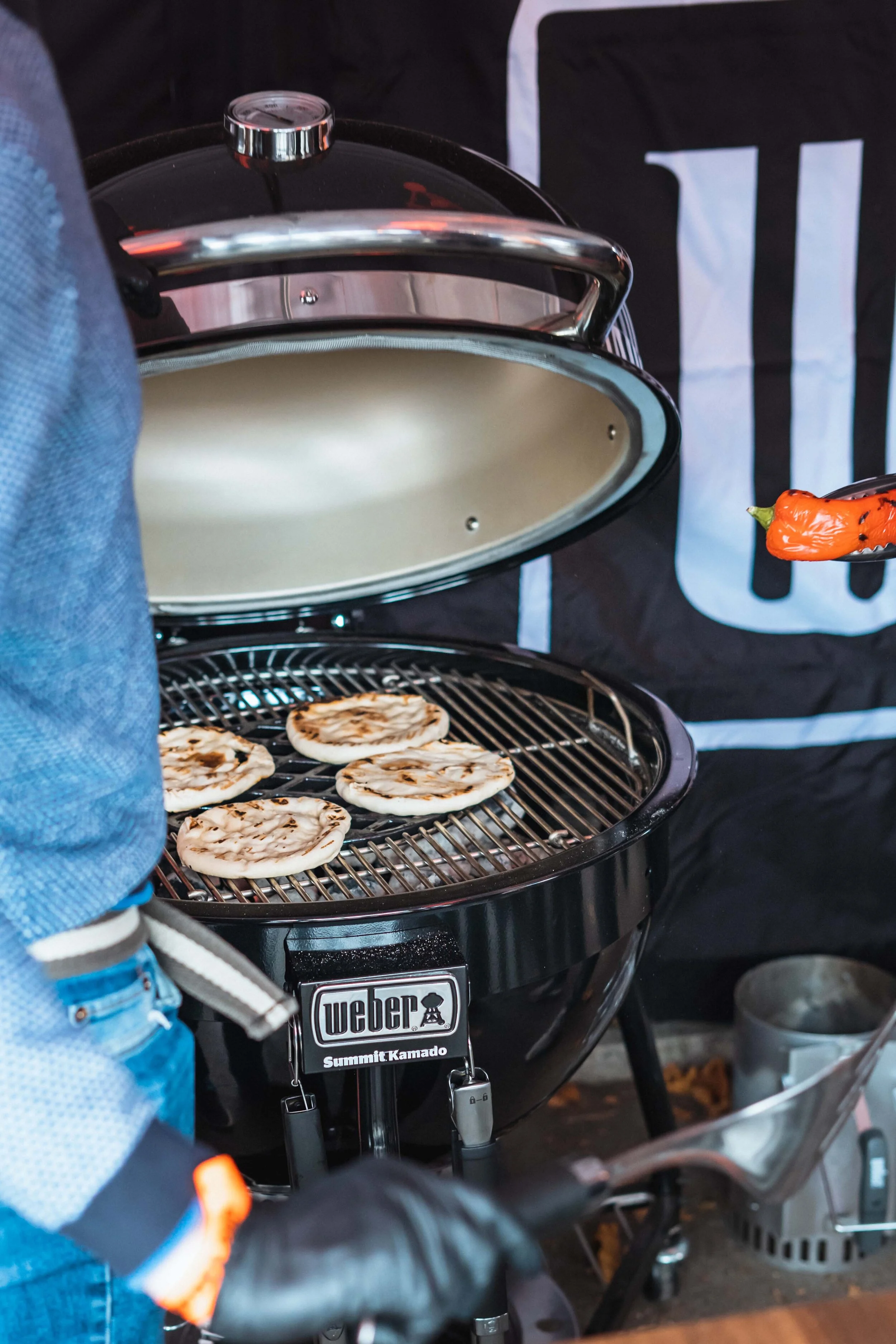 Outdoor grill with round breads cooking on the grates and a person holding a red pepper with tongs.