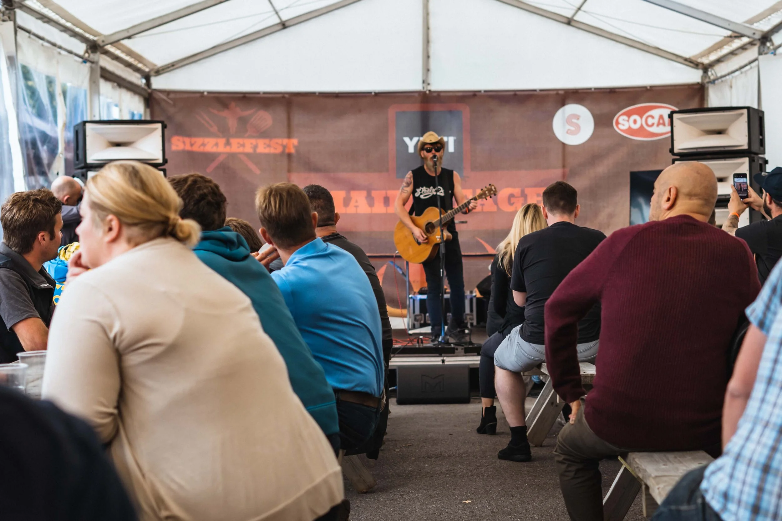 Musician performing with guitar on stage at Sizzlefest event, audience seated on benches in festival tent.