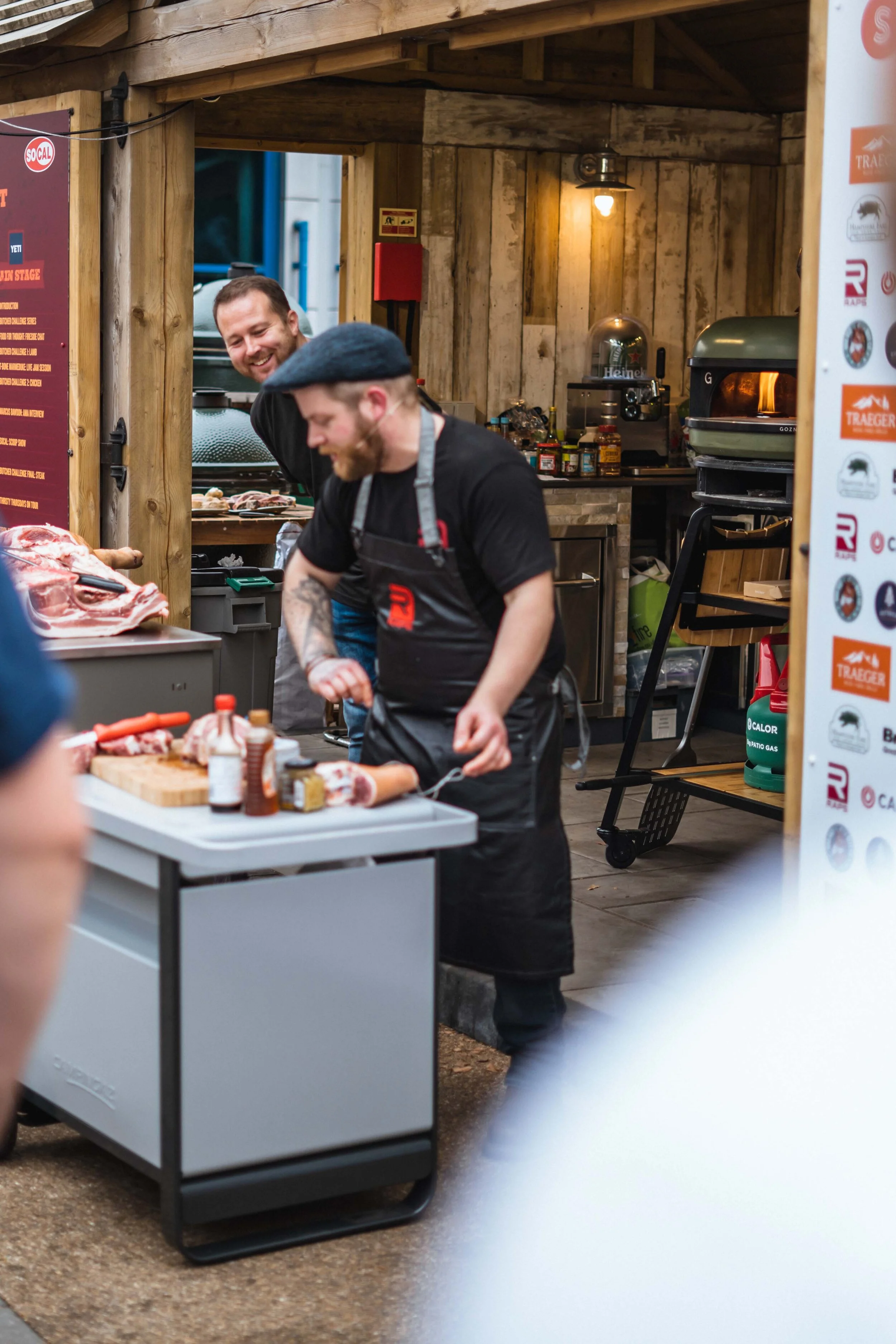 Two men working at an outdoor food stall, one slicing meat on a cutting board, surrounded by cooking supplies and equipment.