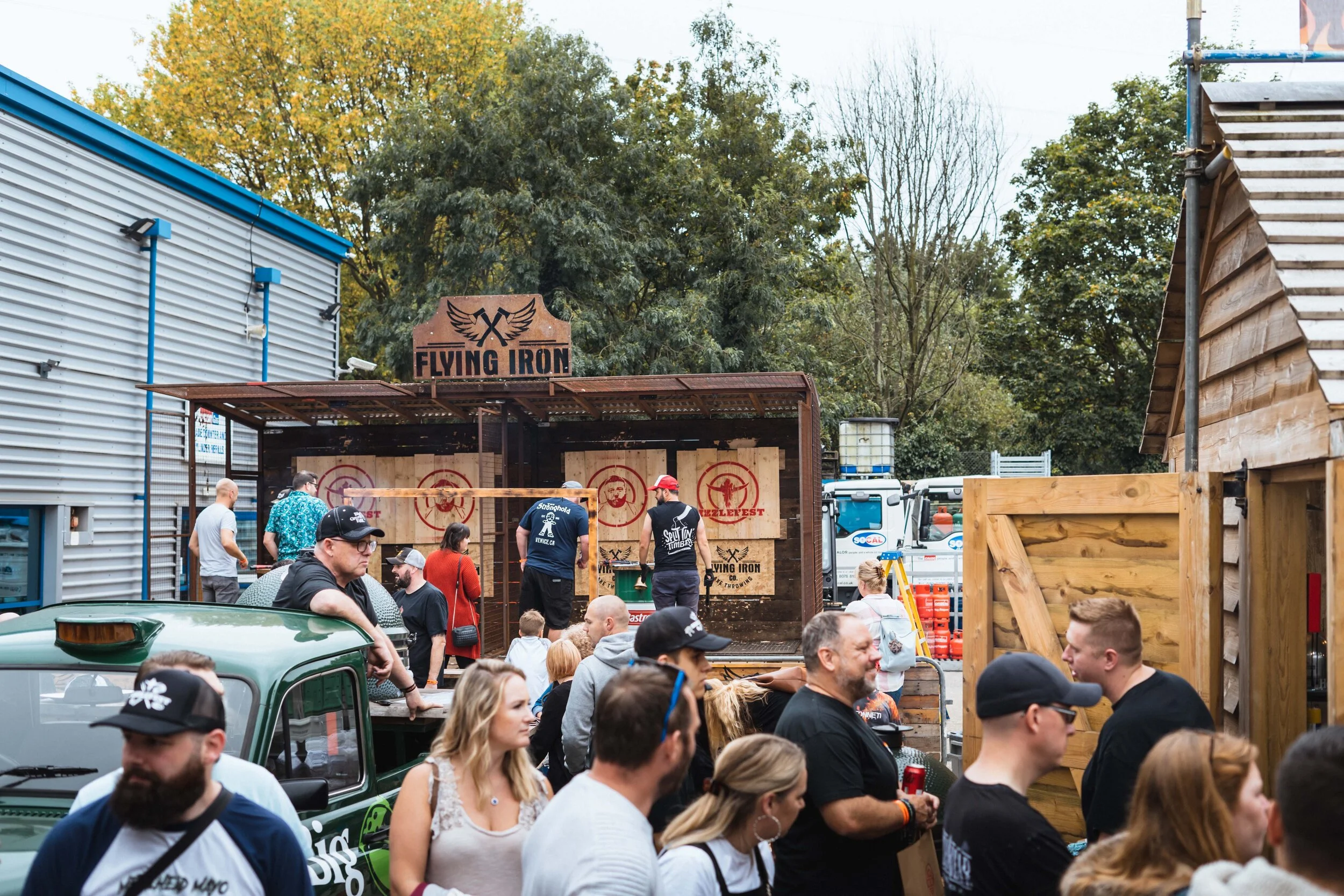 People gathered at an outdoor axe throwing event with targets, hosted by Flying Iron. Various attendees are interacting and observing the activity, surrounded by industrial buildings and trees.
