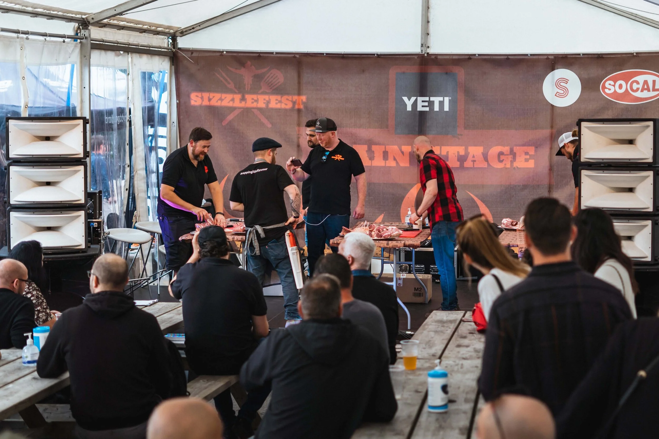 People attending a cooking demonstration or BBQ event under a tent, with speakers and a backdrop displaying logos for Sizzlefest and Yeti.