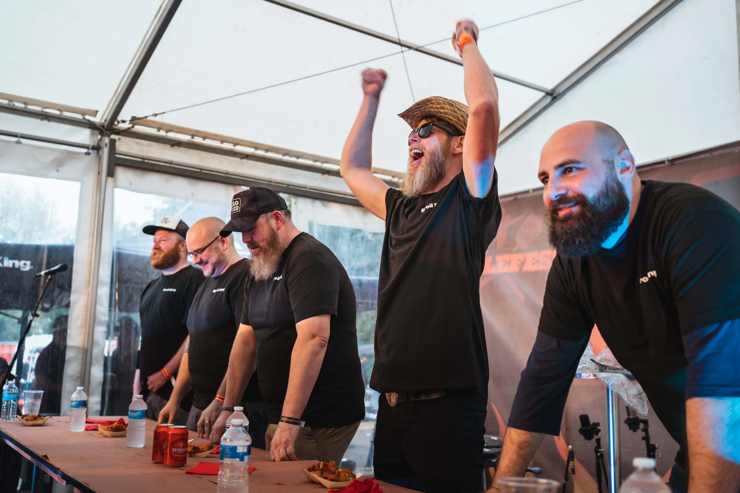Group of men at a food competition, one cheering, under a tent with bottles and plates on a table.