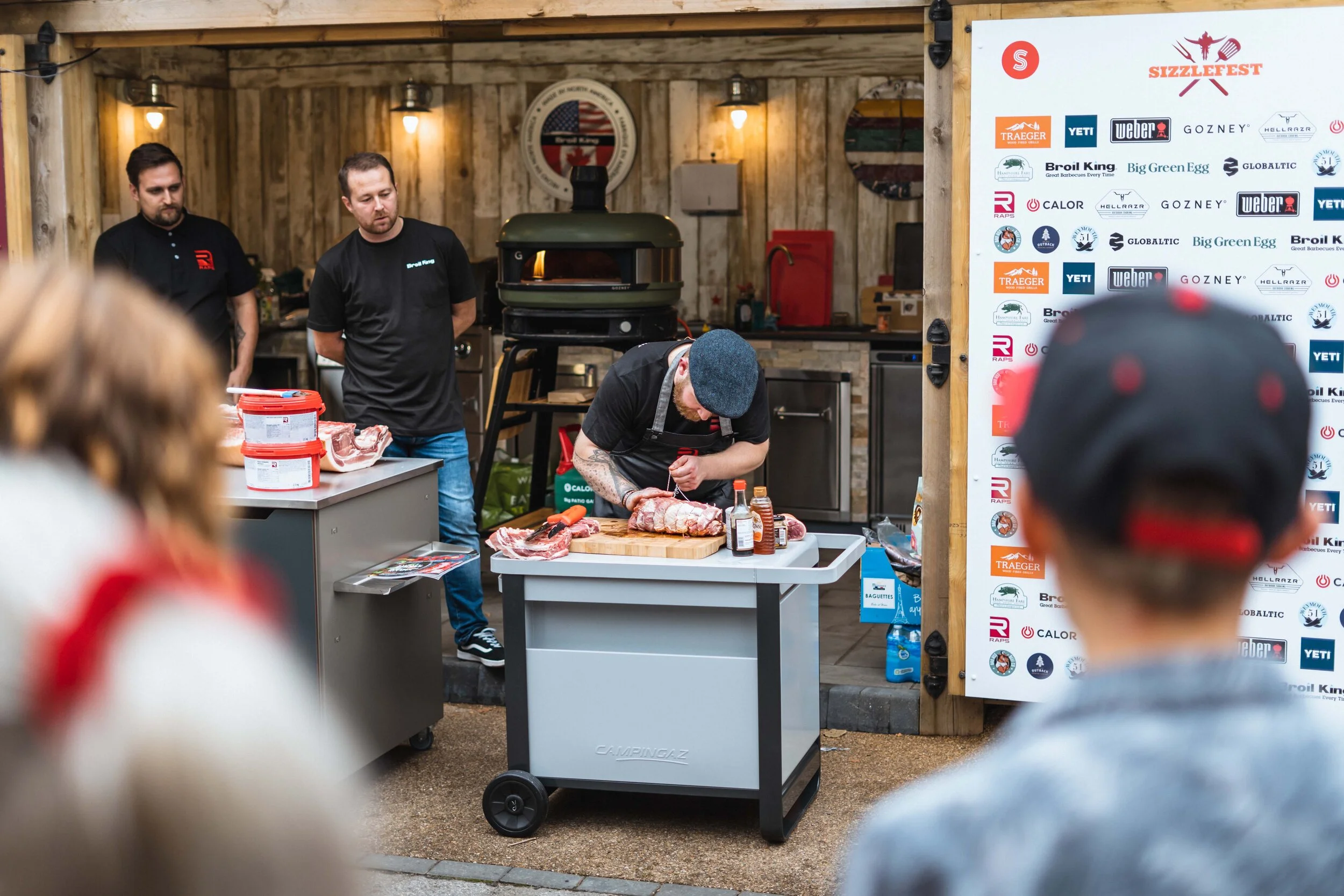 Chef demonstrating meat preparation at Sizzlefest event, various grilling equipment and brand logos visible.