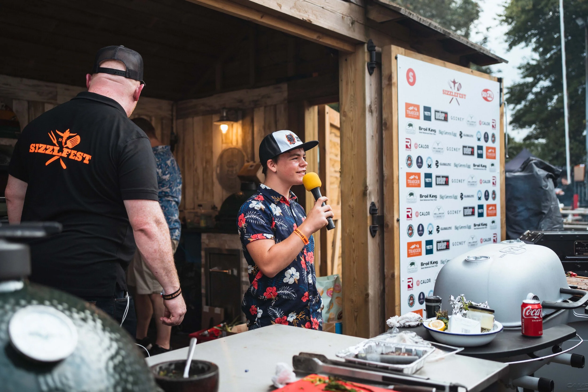 People at a grilling event, one holding a microphone, standing near a table with food and a grill, banner with logos in the background.