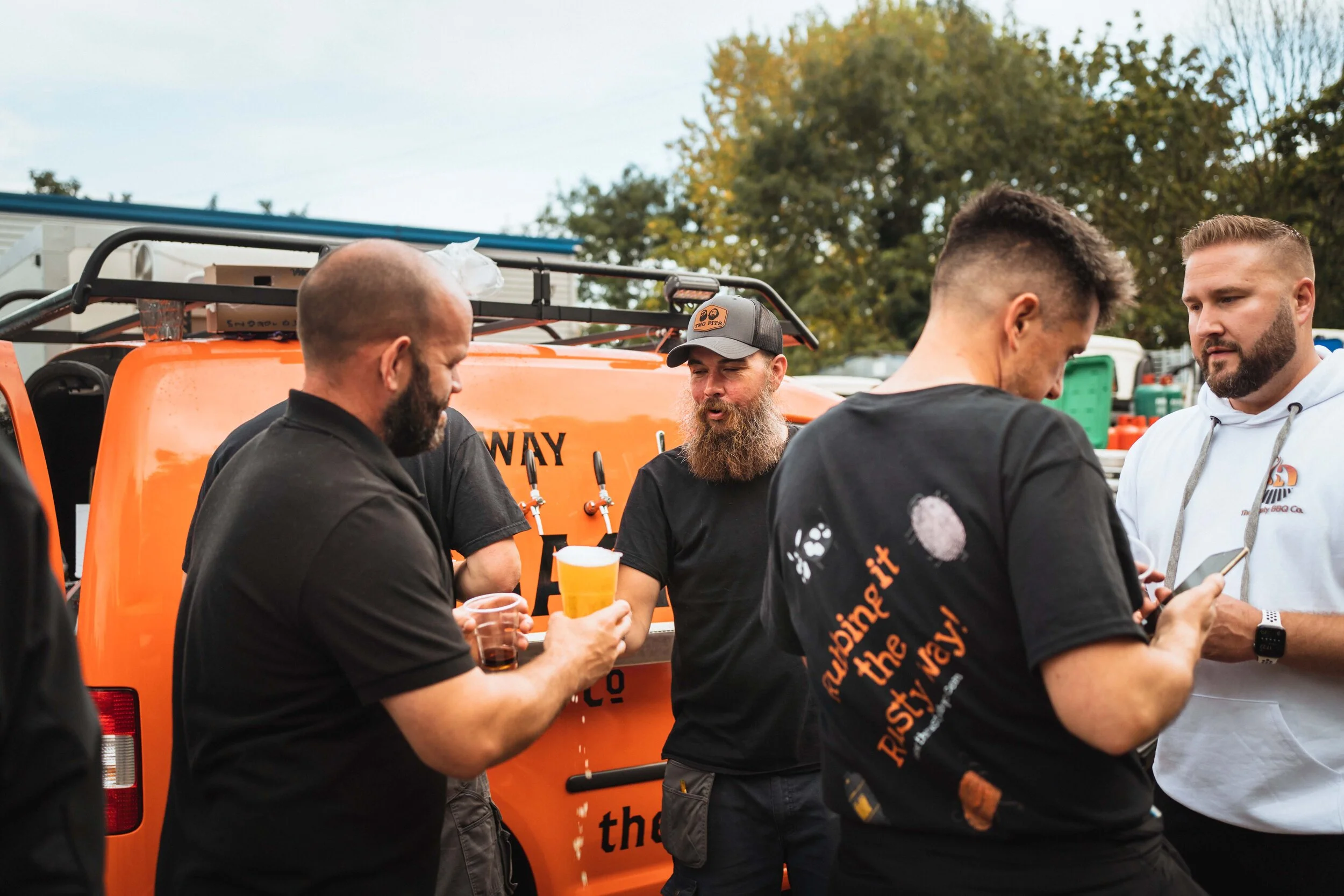 A group of men gathered around an orange vehicle with beer taps in an outdoor setting.