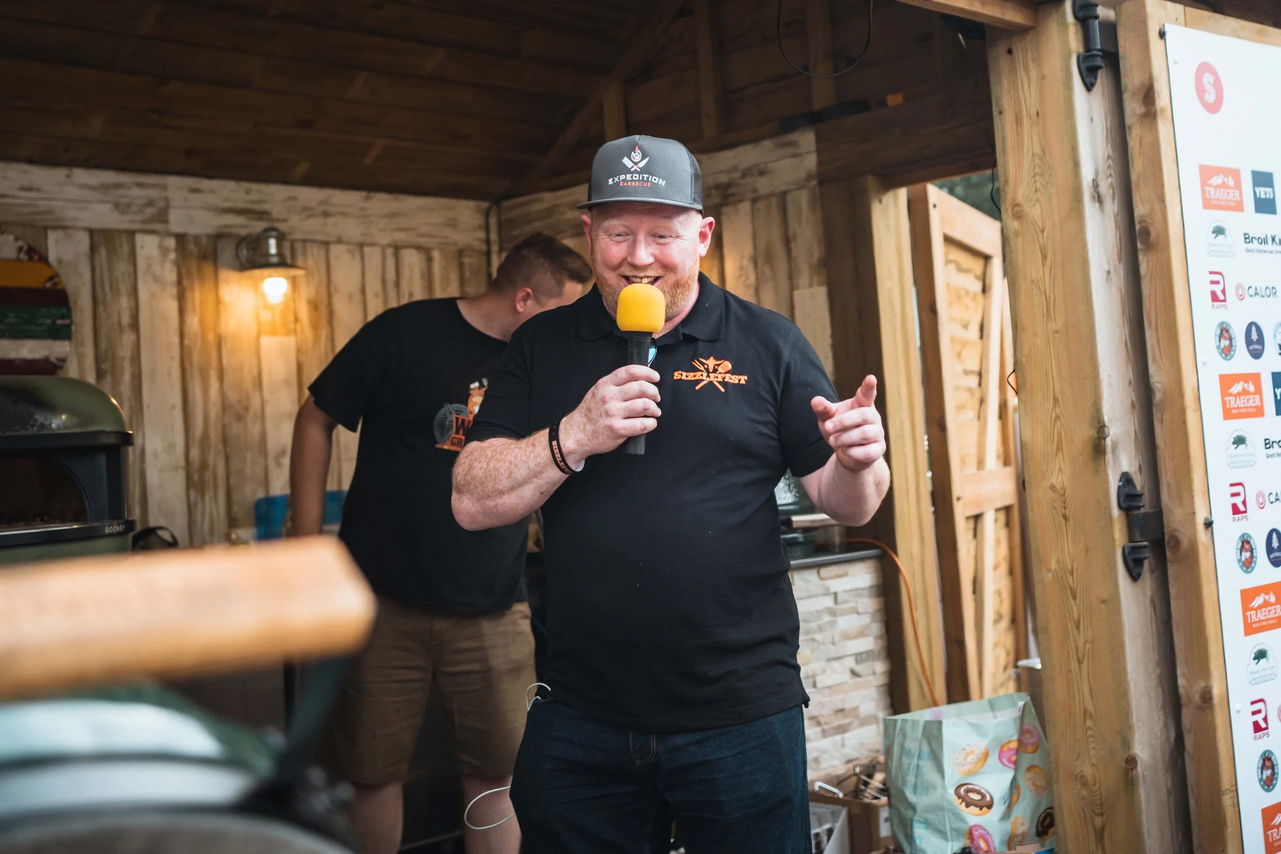 Man with microphone speaking at barbecue event in wooden outdoor kitchen, wearing a cap and black t-shirt with "Sizzlefest" logo.