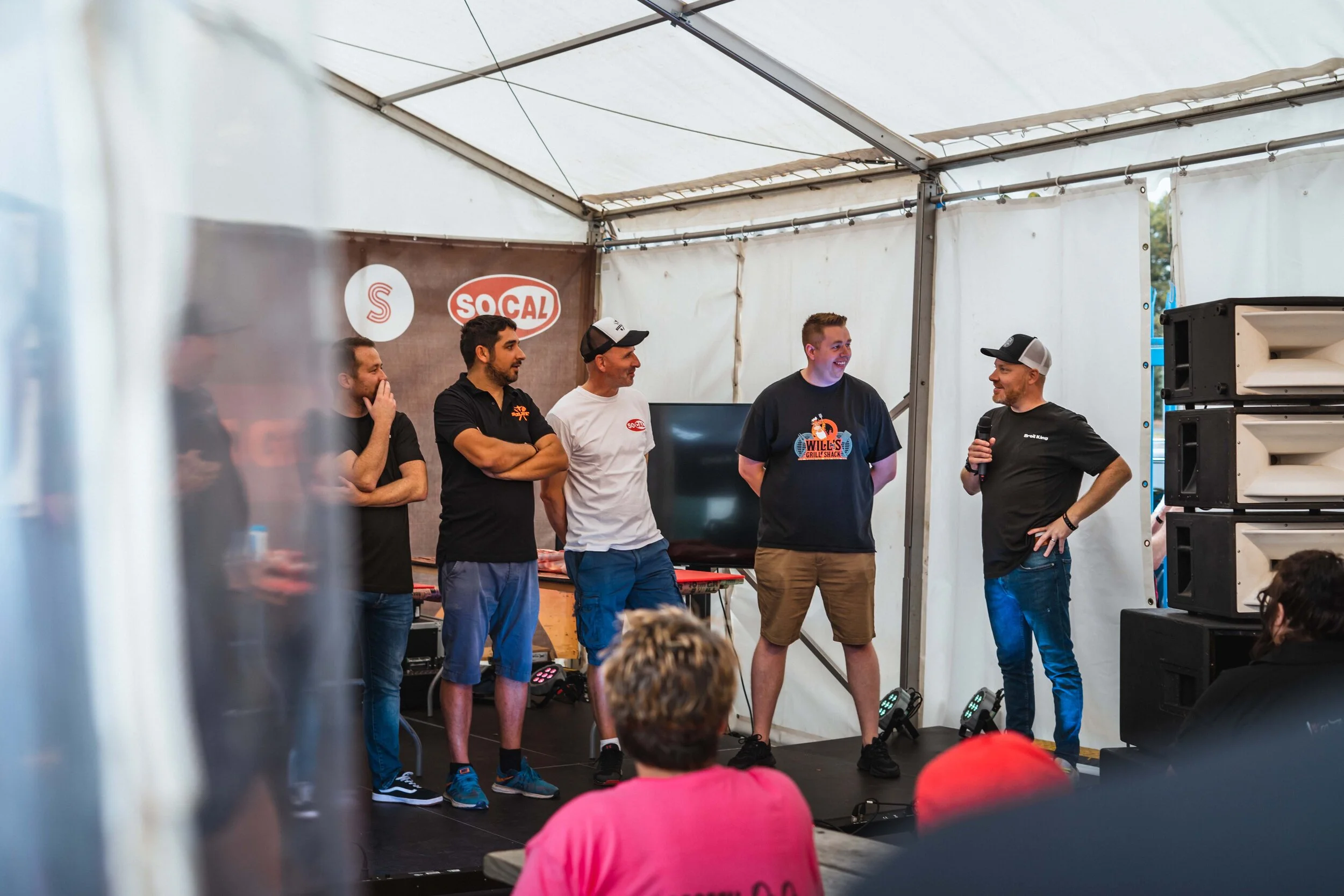 Group of men standing on stage under a tent, one man holding a microphone, audience seated in foreground. Background shows logos and audio equipment.