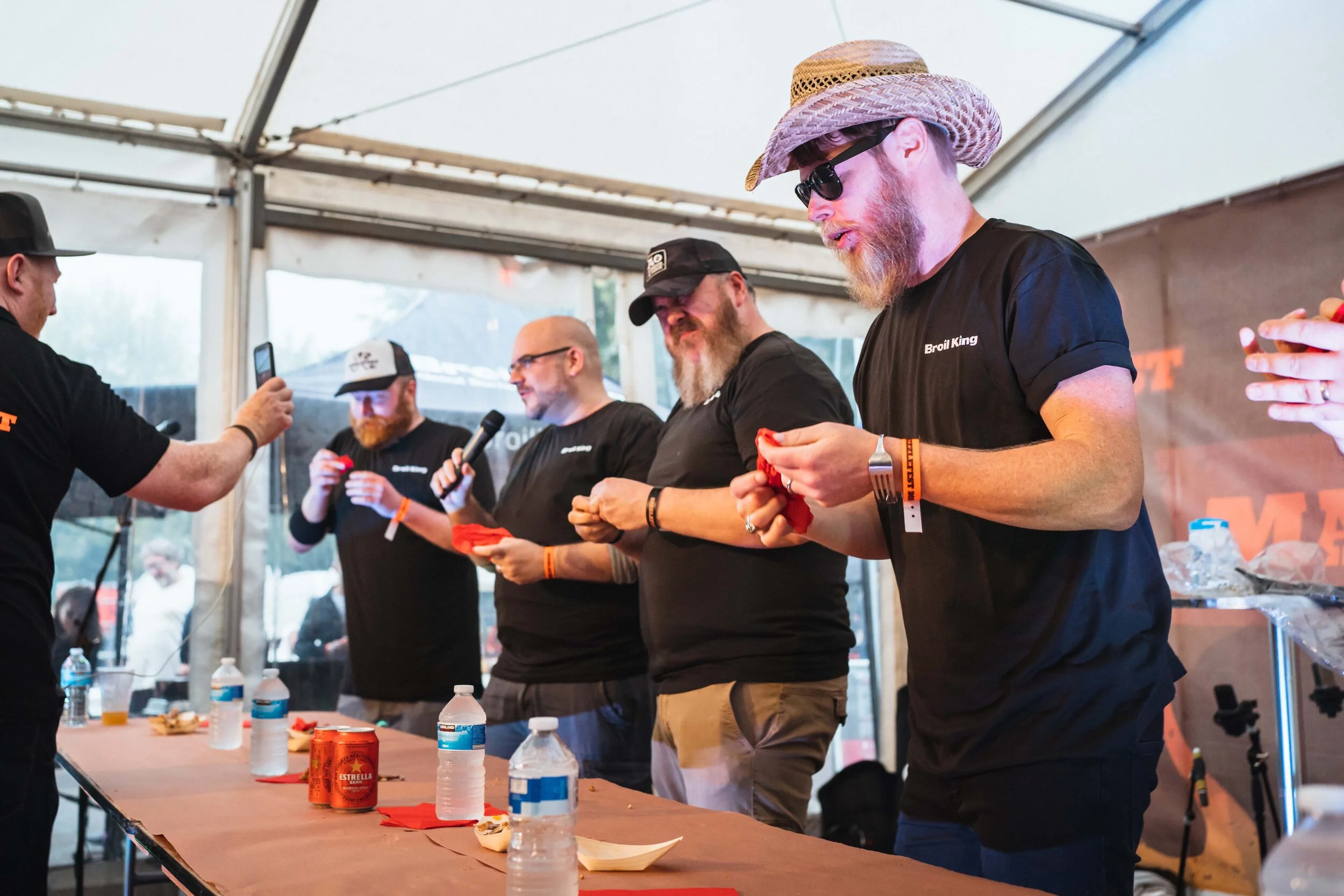 Four men in black shirts participating in a food tasting event indoors, with water bottles on the table and one person holding a microphone.
